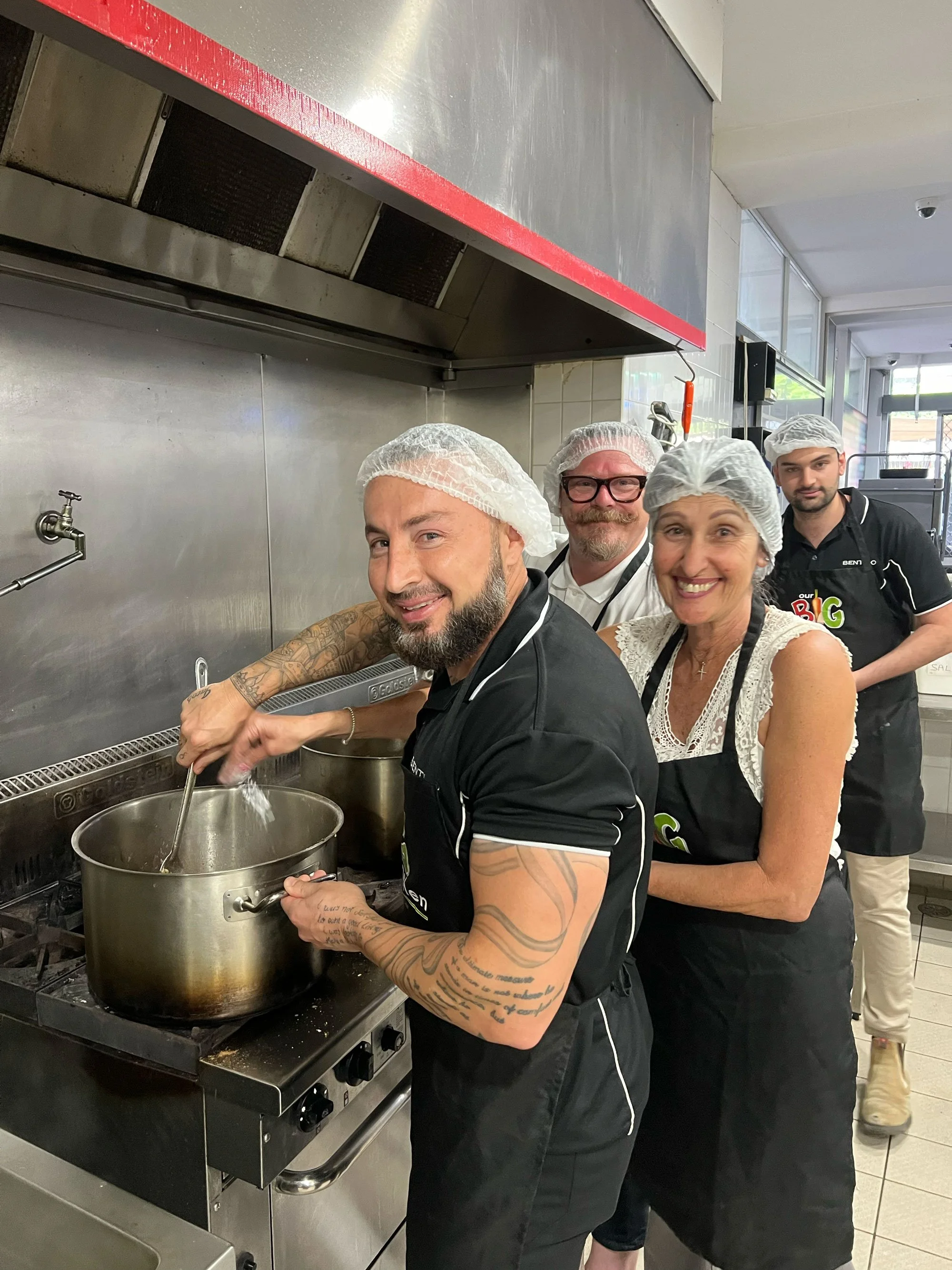 Four people in a commercial kitchen, wearing hairnets and aprons, making soup or sauce in large cooking pots. They are smiling and looking at the camera.