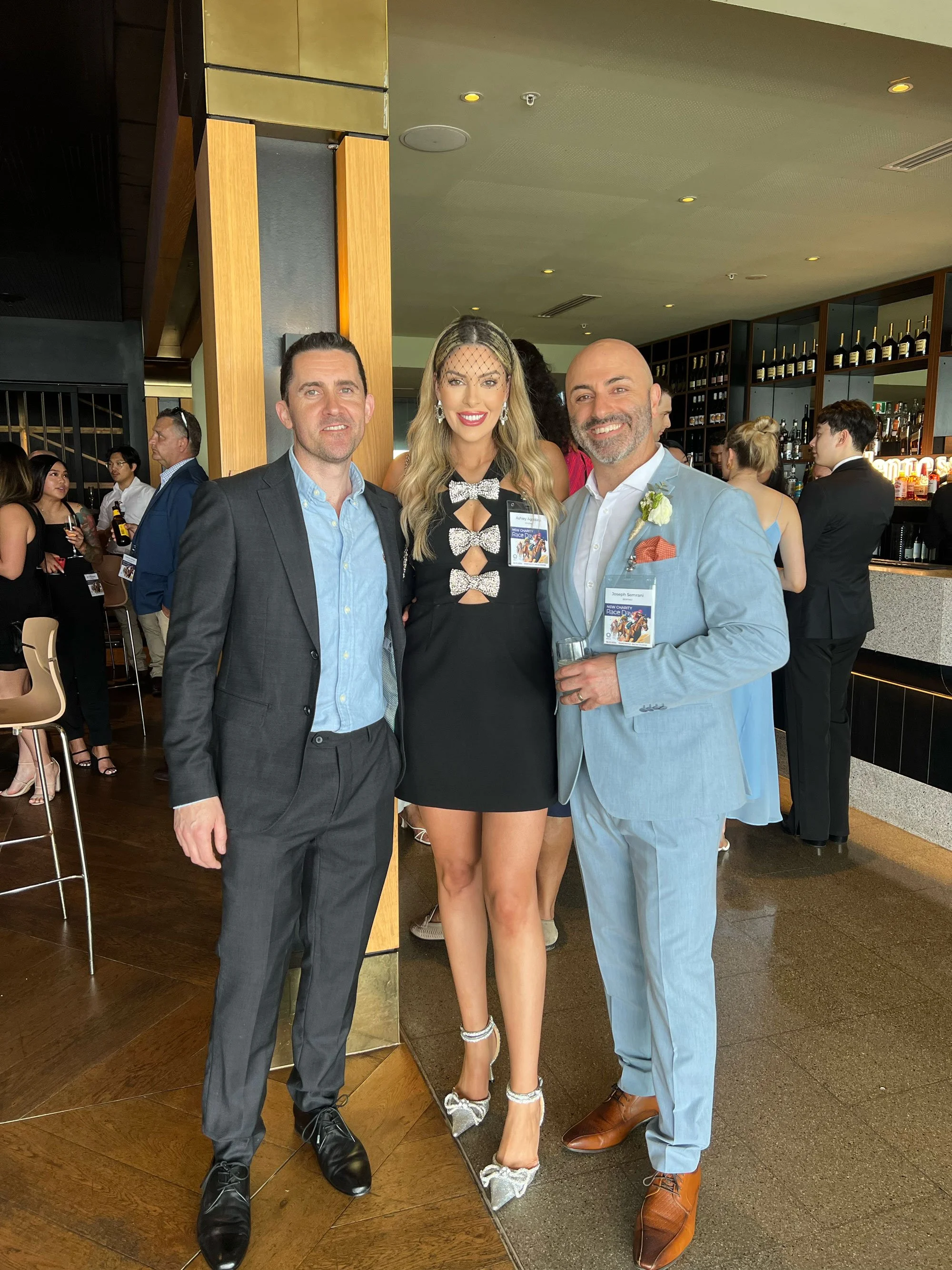 Three people smiling at a social event indoors, with a bar and other guests in the background. The woman in the center is dressed in a black dress with rhinestone bows and silver heels, wearing a veil and earrings. The man on the left is in a gray suit with a blue shirt, and the man on the right is in a light blue suit with a white shirt, a boutonnière, and holding a glass.