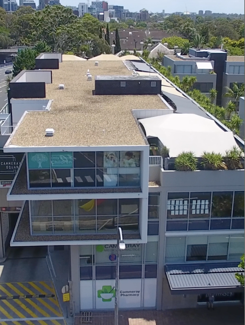 Aerial view of a modern multi-story building with glass windows, greenery on the rooftops, and city skyscrapers in the background. The building includes a pharmacy and medical offices on the ground floor.