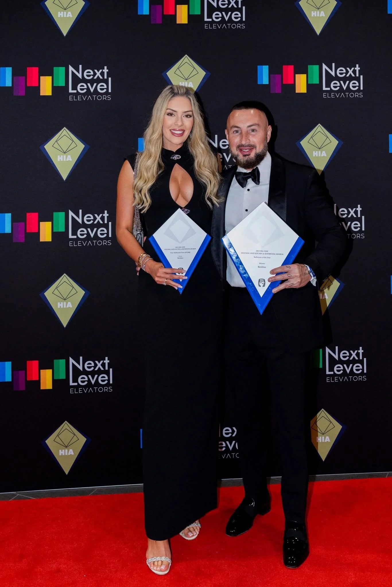 A man and a woman dressed formally, holding awards, standing on a red carpet in front of a black backdrop with the logos 'Next Level Elevators' and 'HIA'. The woman has long blonde hair, is wearing a black dress with a keyhole cutout, and gold heels. The man has short dark hair, a beard, and is wearing a tuxedo with a bow tie and shiny black shoes.