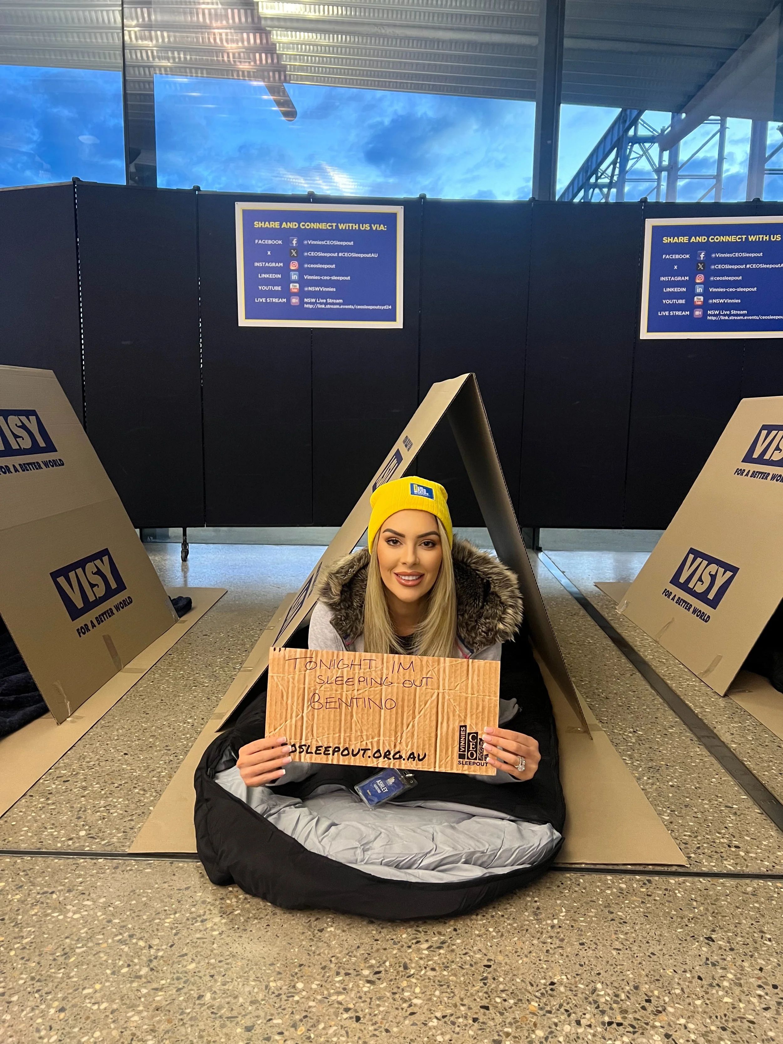 A young woman sitting inside a large black bean bag chair at an airport, holding a handwritten sign that reads 'Tonight I'm sleeping out Bentino' with a website link and event hashtag. She is wearing a yellow beanie and has long blonde hair, and is smiling at the camera. The background shows a dark wall with informational signs and a large window showing the sky outside.