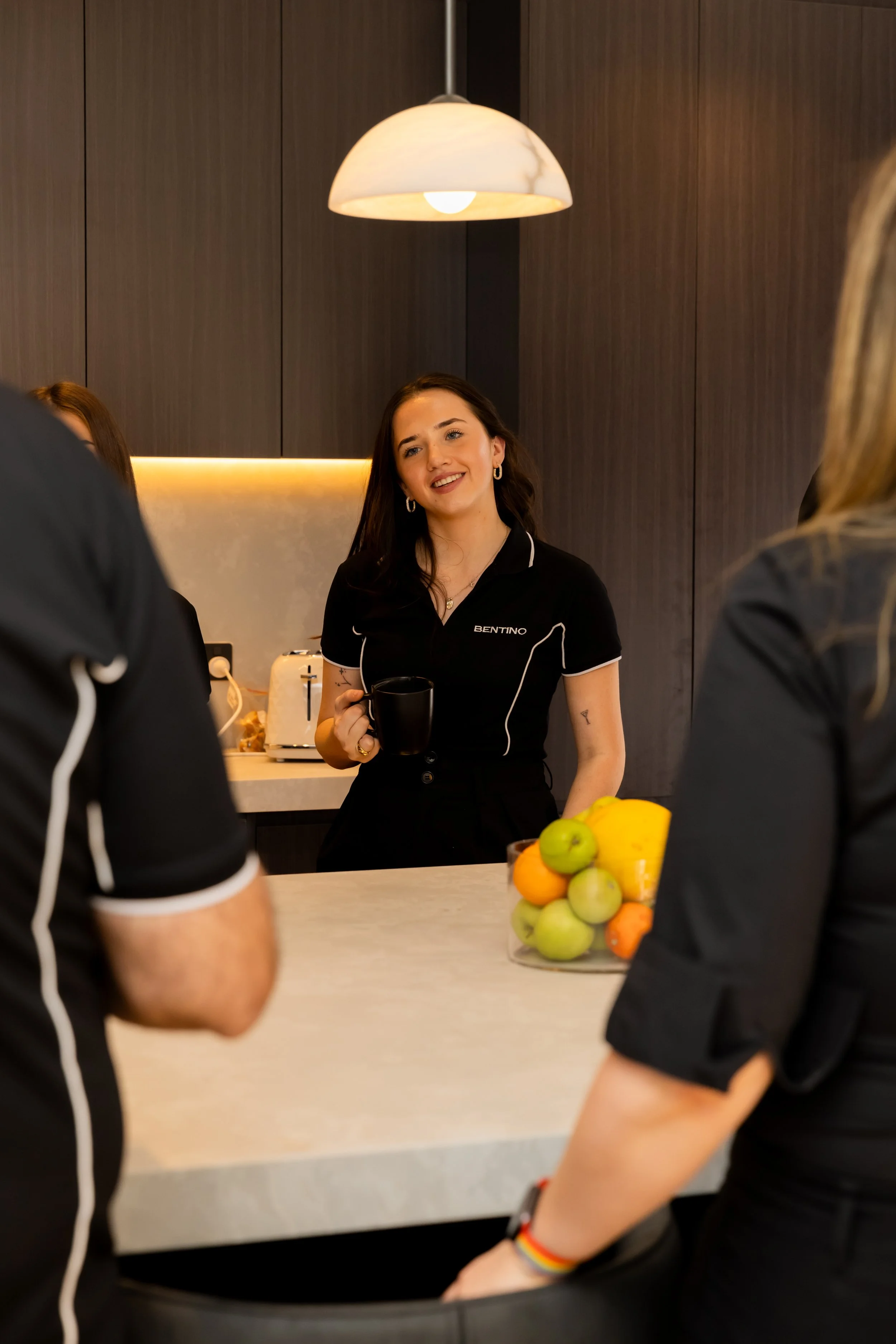 A woman in a black uniform with the name 'BENTINO' on it, smiling and holding a mug, standing in a kitchen with dark wood cabinets and a lit light fixture above. There are other people around, partially visible, and a bowl of various fruits on the counter.