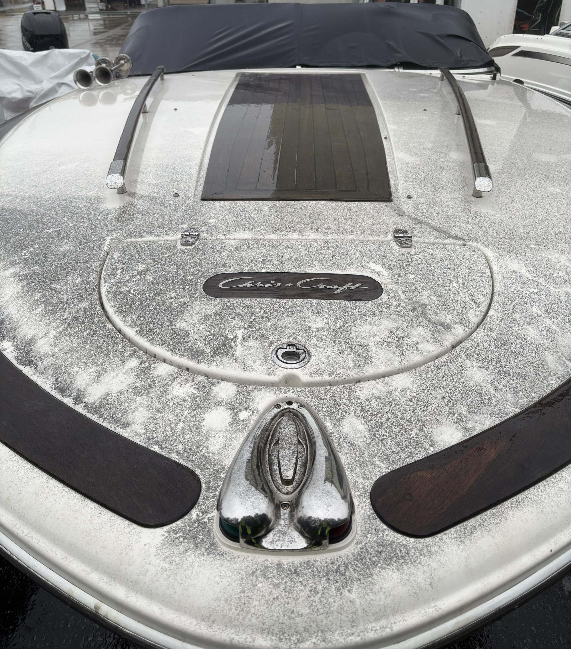 The front of a Chris-Craft boat with a shiny, metallic surface that appears weathered and aged, with some water droplets. The boat has a solar panel mounted on the top, two metal handrails, and a component resembling a rider or seat with a reflective surface. There are also some small vents, and the boat's nameplate can be seen at the bottom, reading 'Chris-Craft'.