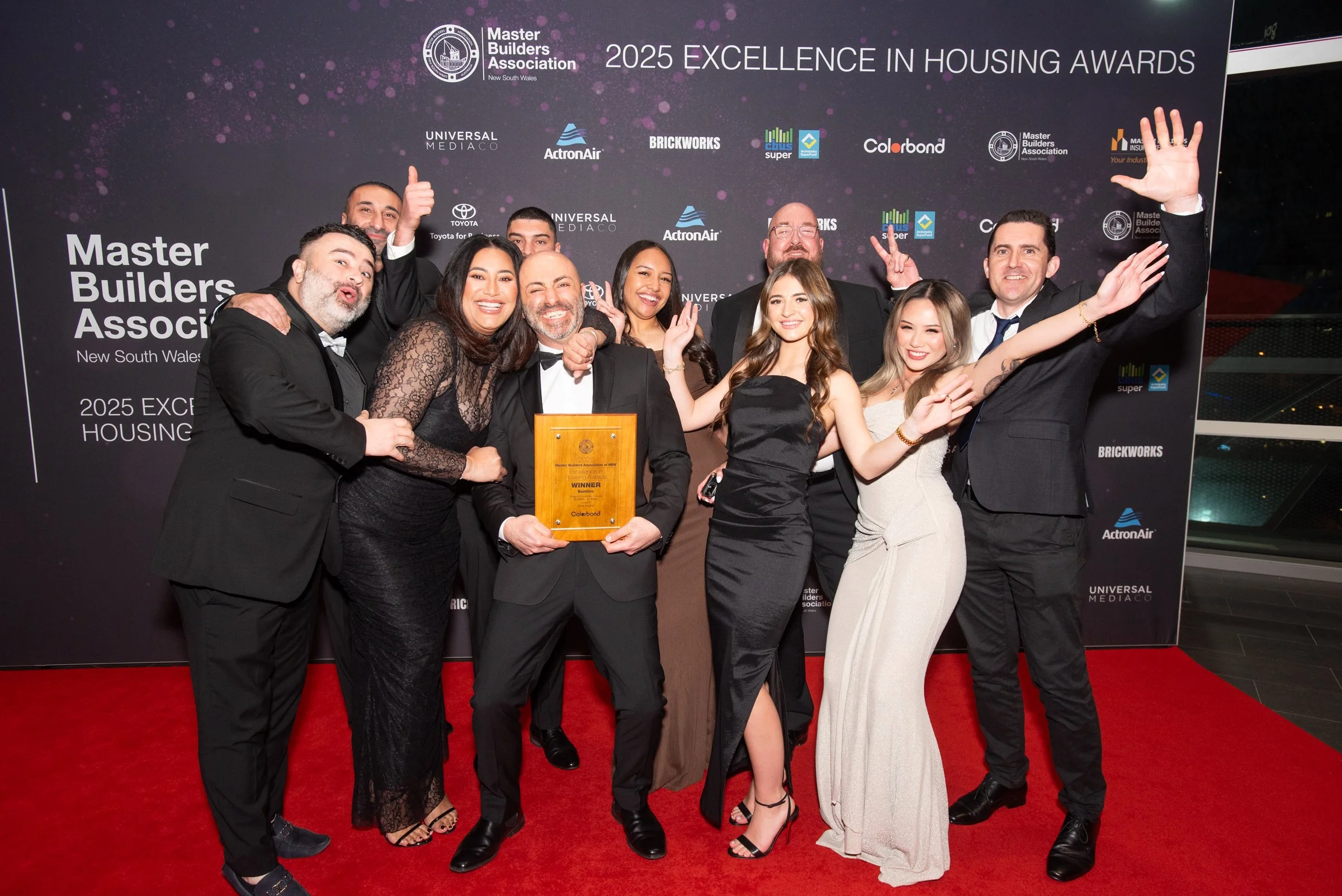 A group of people in formal attire celebrating at an awards event. They are smiling, some with arms around each other, and one person is holding a wooden plaque. A man at the front has a fist raised, and another is raising a hand with fingers spread. The background shows a black backdrop with event logos and text, including "Master Builders Association" and "2025 Excellence in Housing Awards." The event appears to be held indoors, with a red carpet beneath their feet.