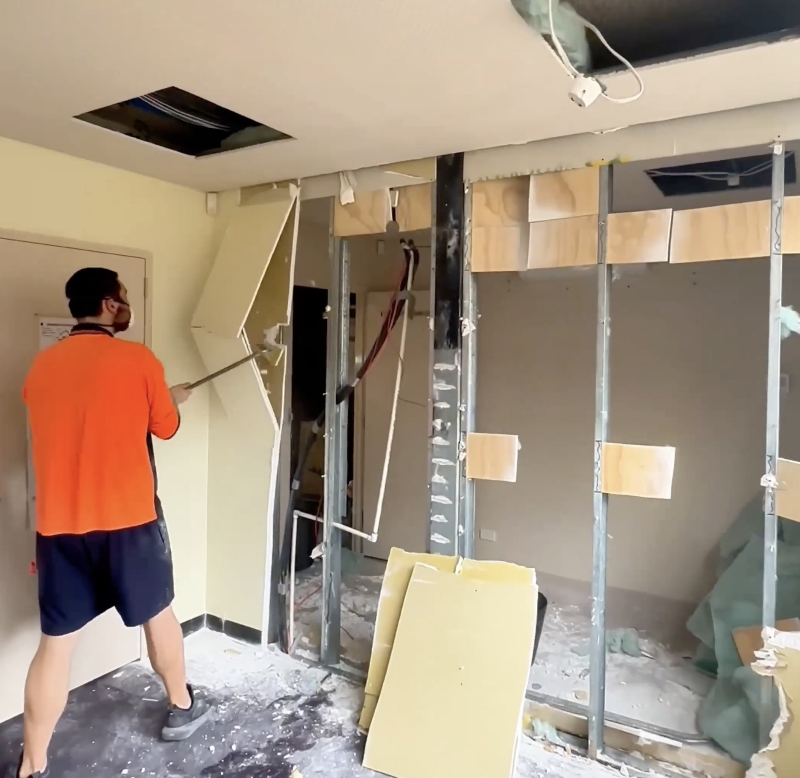 Construction worker demolishing interior wall with a crowbar, exposed metal studs, drywall debris on the floor, and ceiling opening.