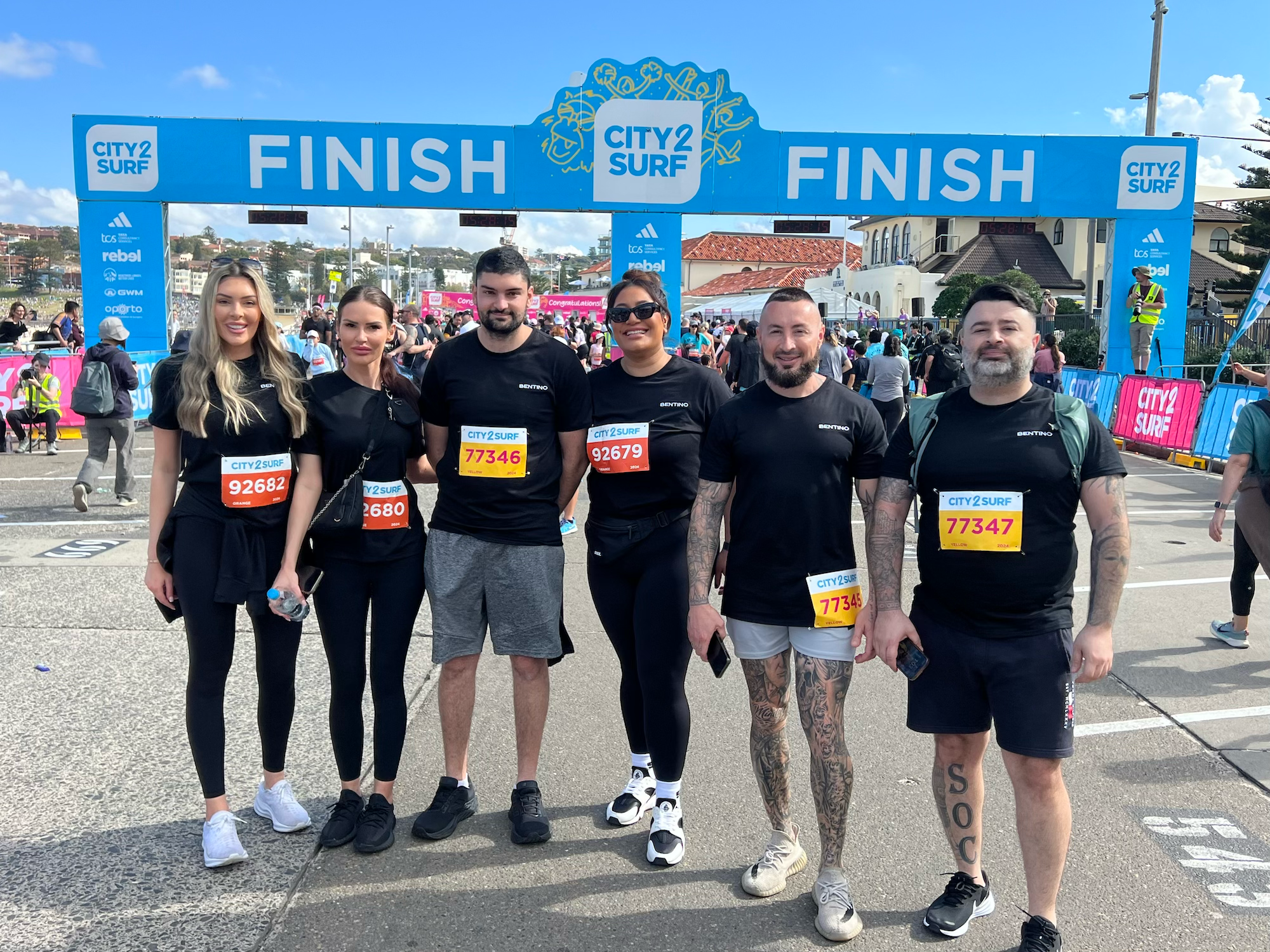 Group of five people standing in front of the finish line at a city surf event, smiling and wearing race bibs.