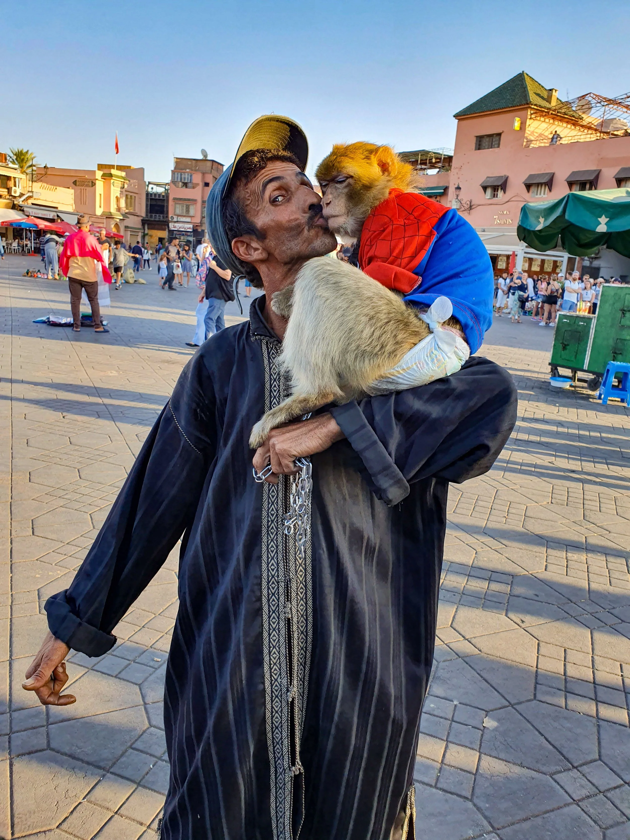 A man and a monkey sharing a kiss in a busy square with buildings and people in the background.