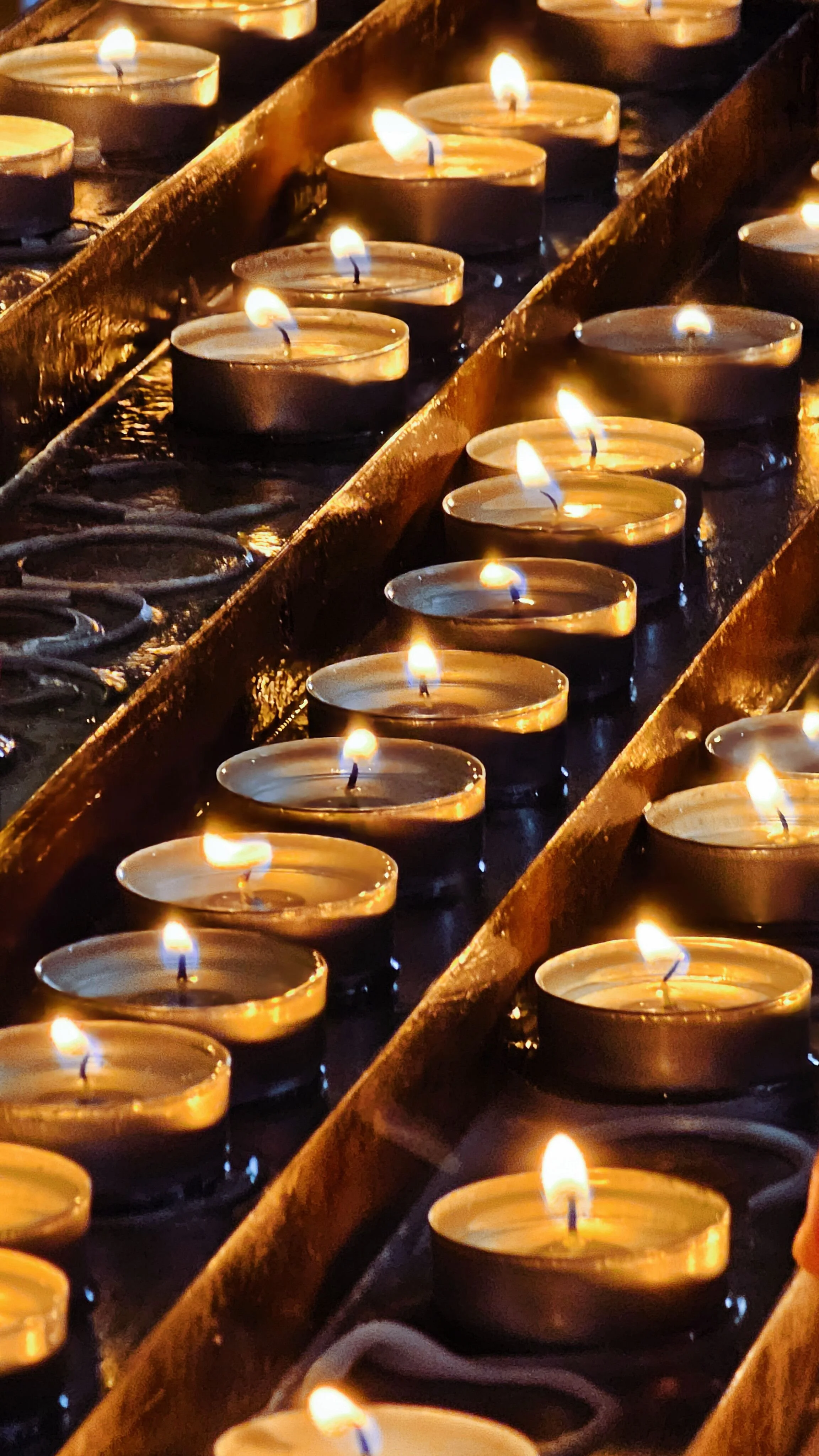Multiple lit tealight candles arranged in rows inside a reflective container with a warm, flickering glow.