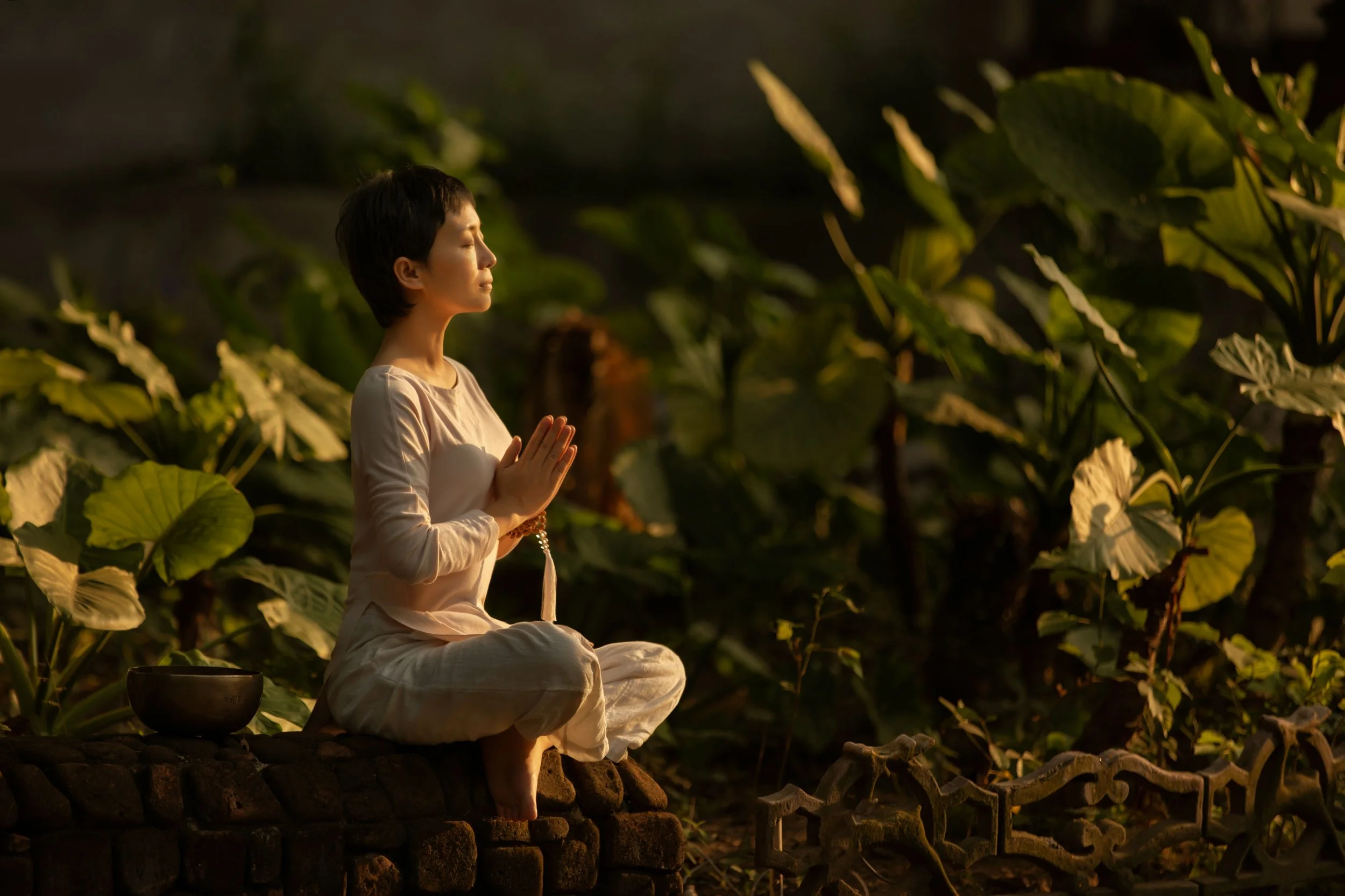 A person dressed in white traditional clothing meditating outdoors at sunset, sitting cross-legged on a stone ledge with hands in prayer position and a prayer bead necklace, surrounded by large green leaves.