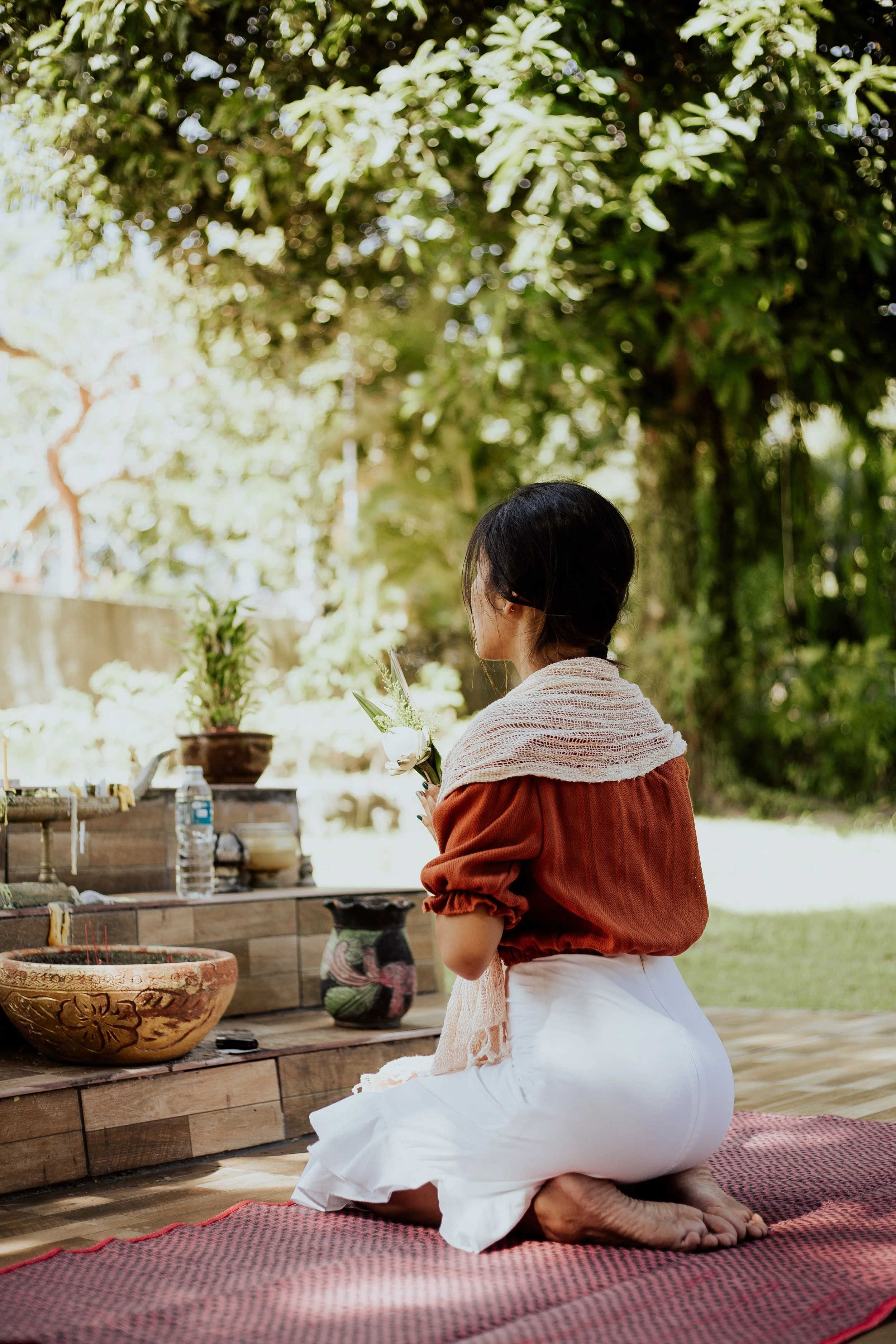 A woman with short dark hair kneels on a red and black woven mat outdoors, holding white flowers and facing an altar with potted plants, bowls, and incense. She is wearing a rust-colored top with puffed sleeves and a white skirt, with her head bowed.