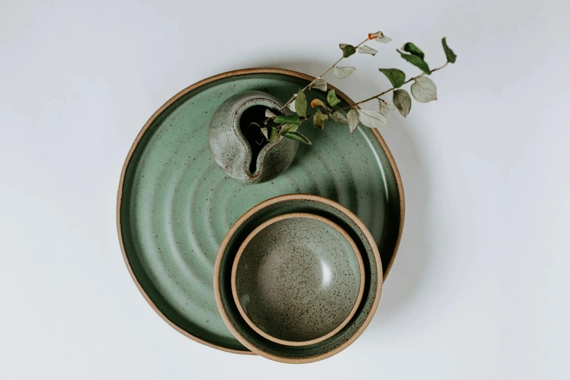 Stacked green and beige ceramic bowls with a small ceramic vase containing dried leaves on a green tray, all placed on a white surface.