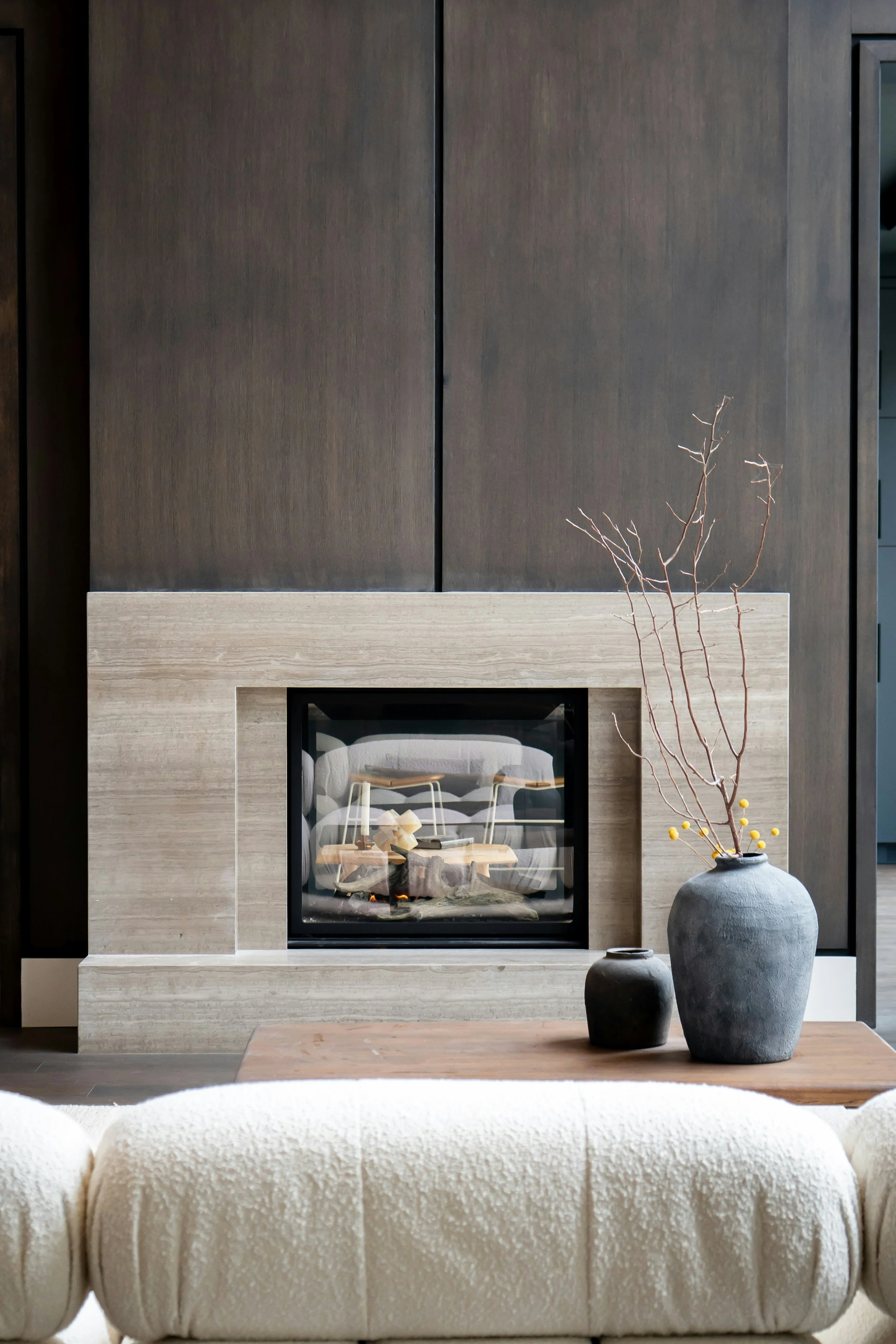 Interior scene featuring a modern fireplace with a wooden mantel, two vases with thin, leafless branches, and a plush cream-colored sofa in the foreground.
