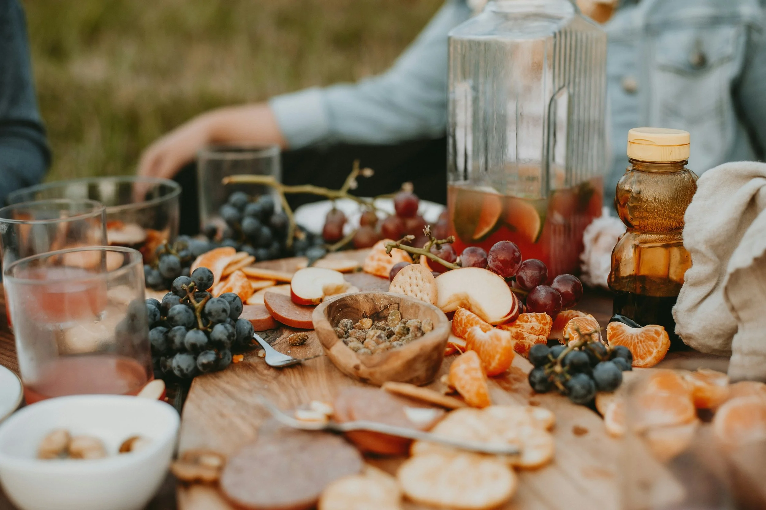 A picnic table set with grapes, sliced fruits, cheese, nuts, crackers, and honey jars, outdoors on a wooden surface with a blurred background.
