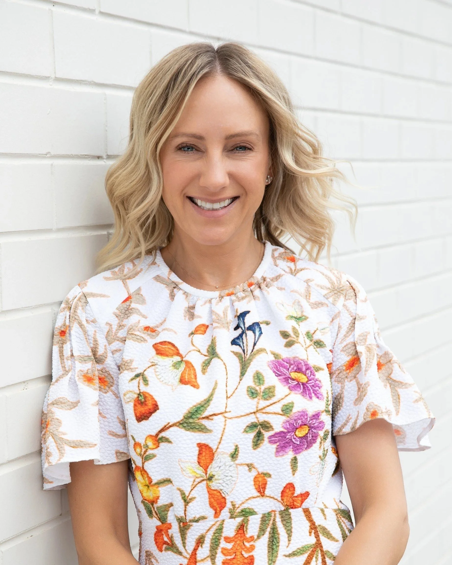 A woman with blonde, wavy hair and blue eyes smiling in front of a white brick wall, wearing a white dress with colorful floral embroidery.