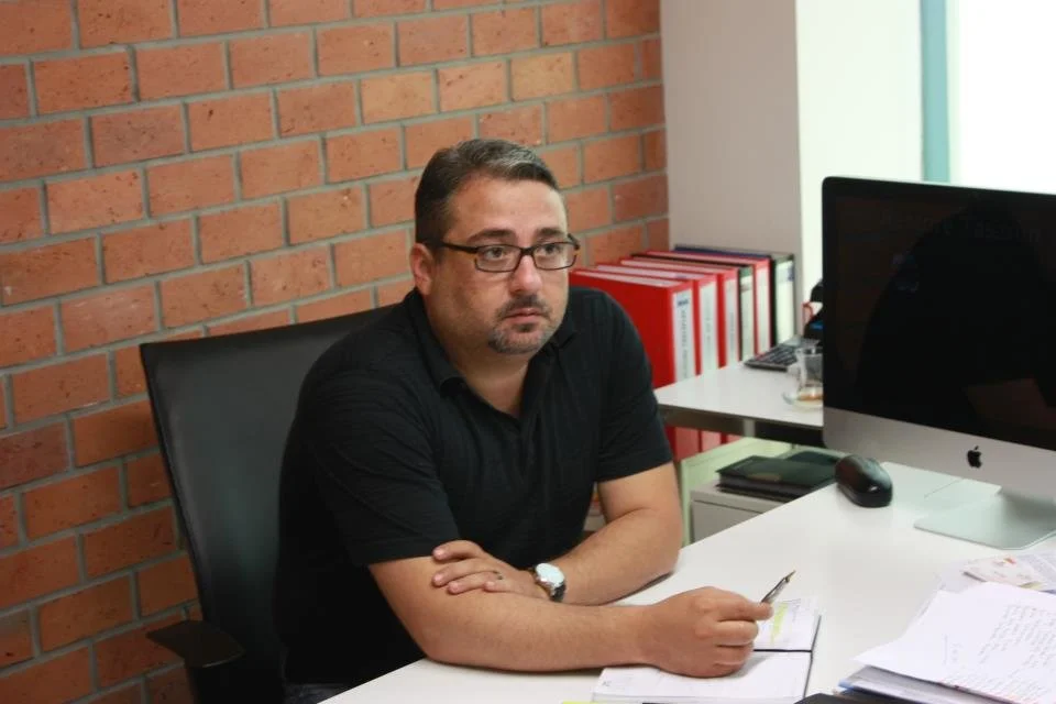 Ofis ortamında, masasında notlar tutan, bilgisayar ve düzenli dosya rafları olan bir adam.