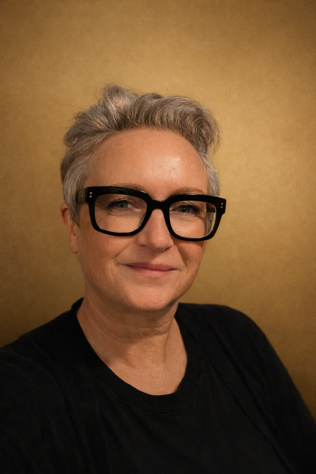 A woman with short gray hair and black glasses smiling at the camera against a beige wall.