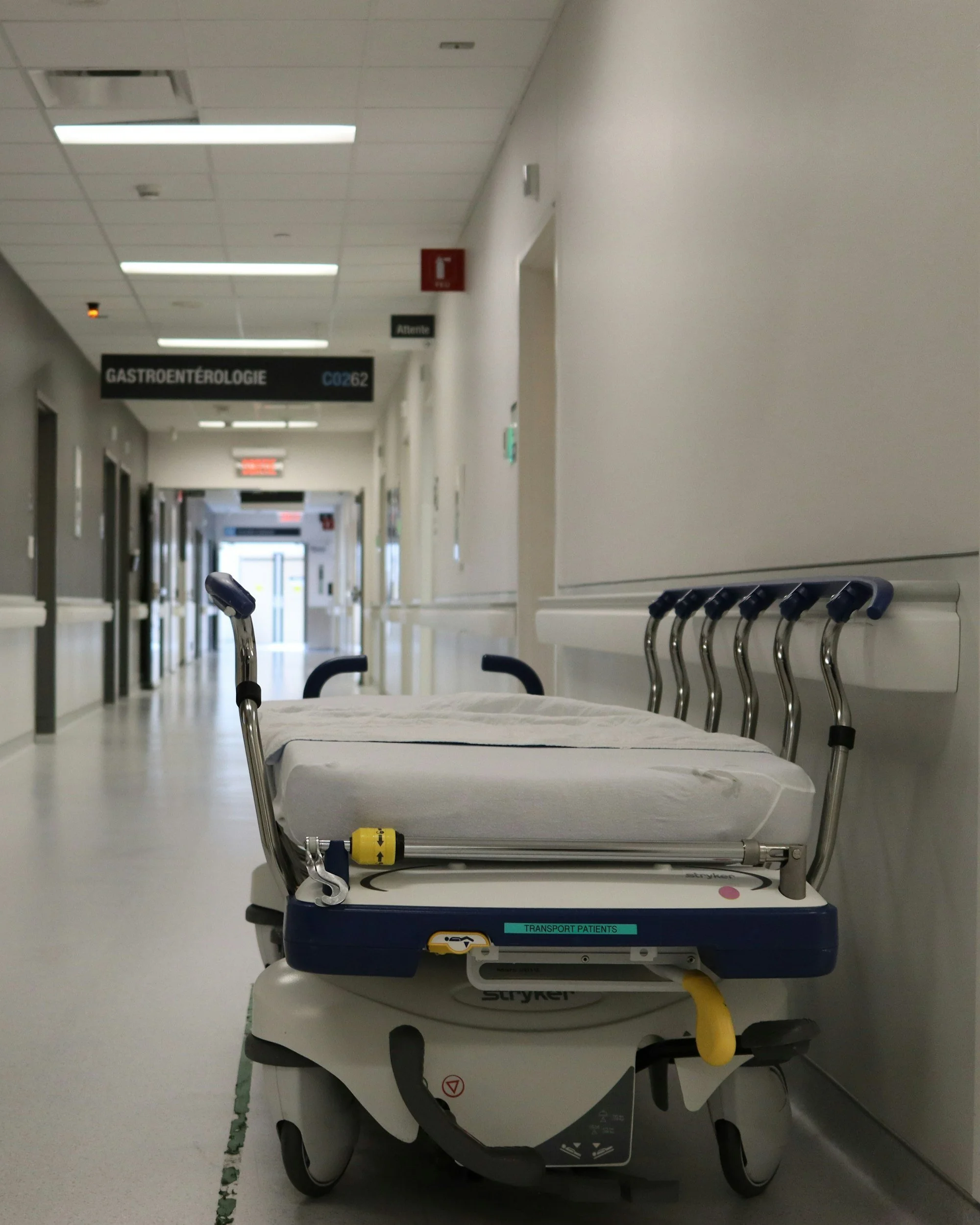 Hospital hallway with an empty patient transport bed in the foreground and signs hanging from the ceiling indicating a gastroenterology department and a fire extinguisher.