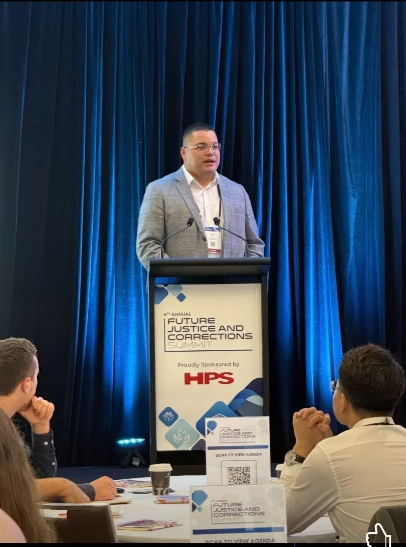 A man in a gray blazer and glasses standing at a podium speaking at the 9th Annual Future Justice and Corrections Summit. The podium has a sign with event details and sponsor HPS, with a QR code for viewing the agenda. Audience members seated in front are listening.