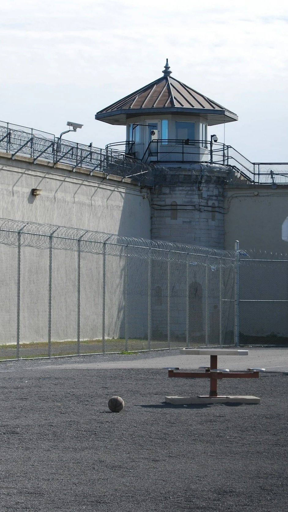 View of a tall prison or correctional facility's watchtower with barbed wire fencing and a basketball on the ground.