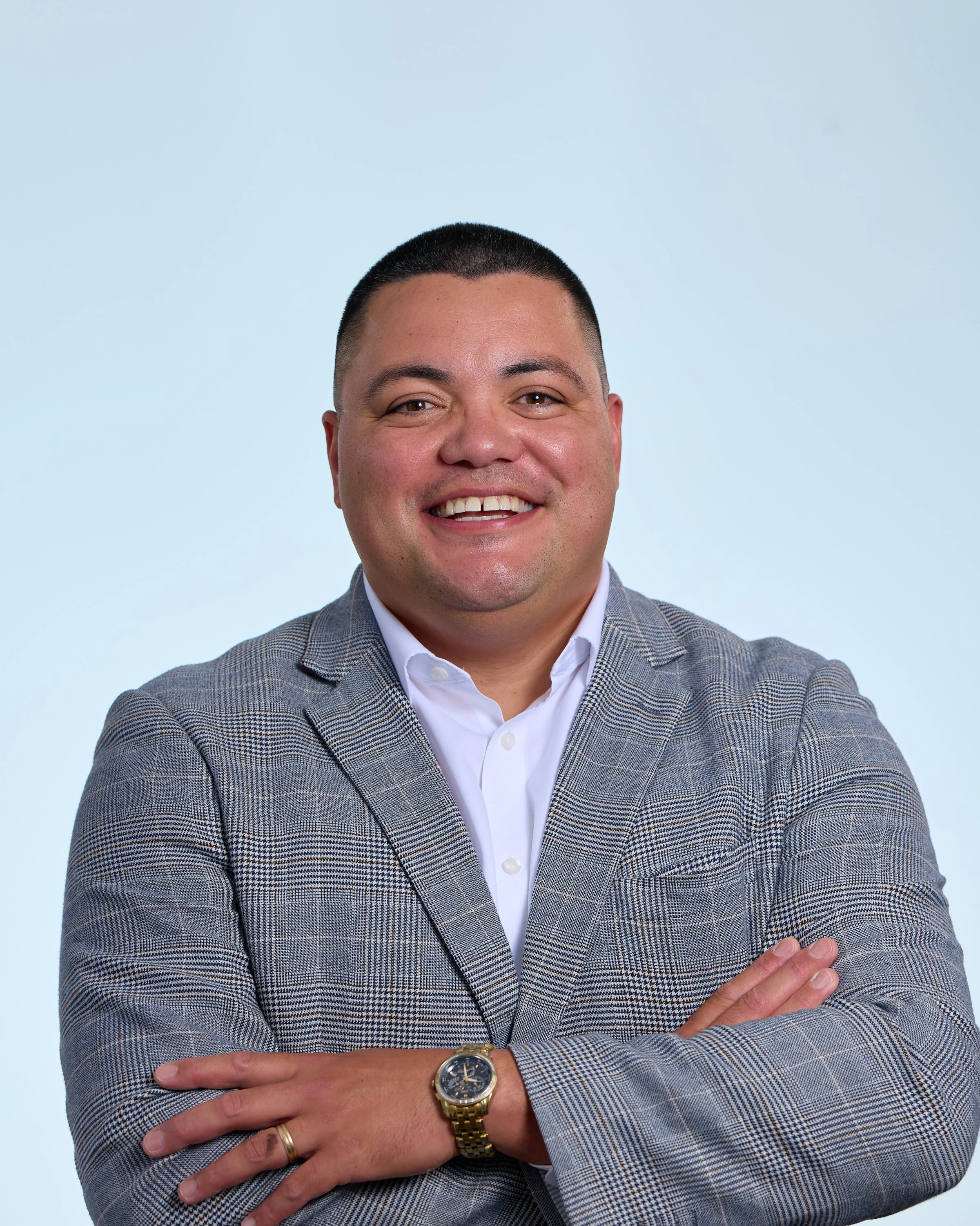 Close-up of a smiling man wearing a gray plaid blazer, white shirt, gold watch, and wedding ring, with arms crossed in front of a light blue background.