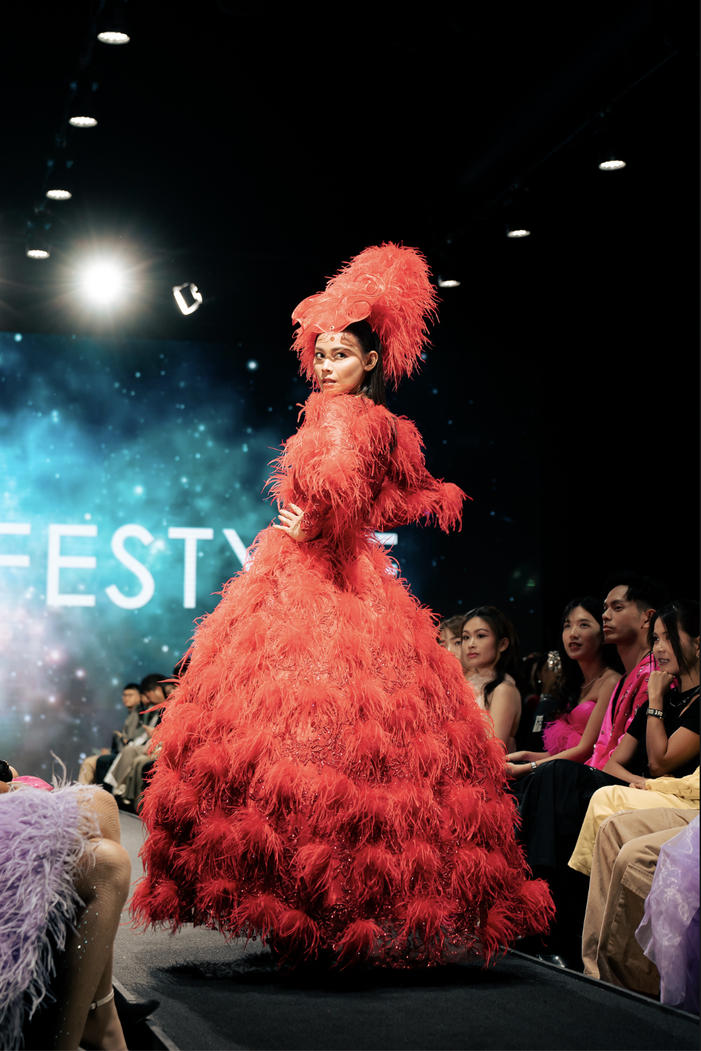 A model walking on the runway wearing a vibrant red feathered gown and a matching headpiece inspired by a bird, during a fashion show.