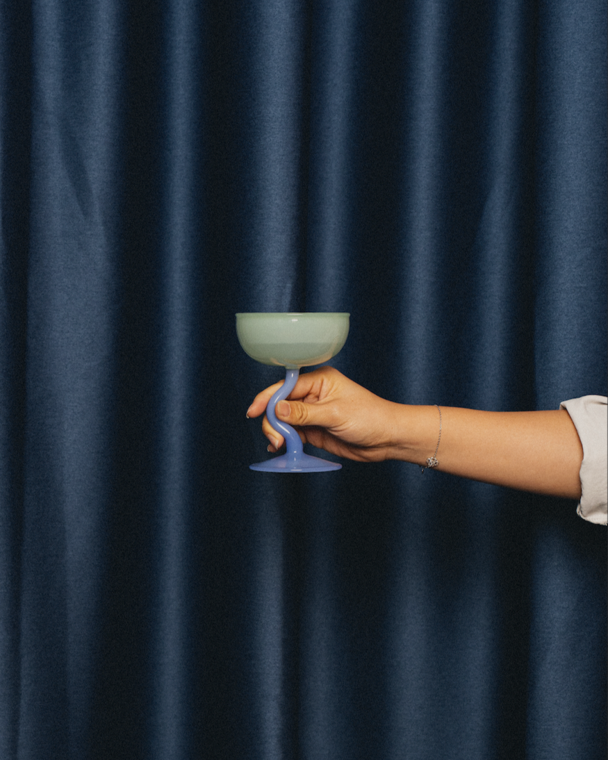 A hand holding a ceramic goblet with a blue twisted stem against a dark blue curtain background.