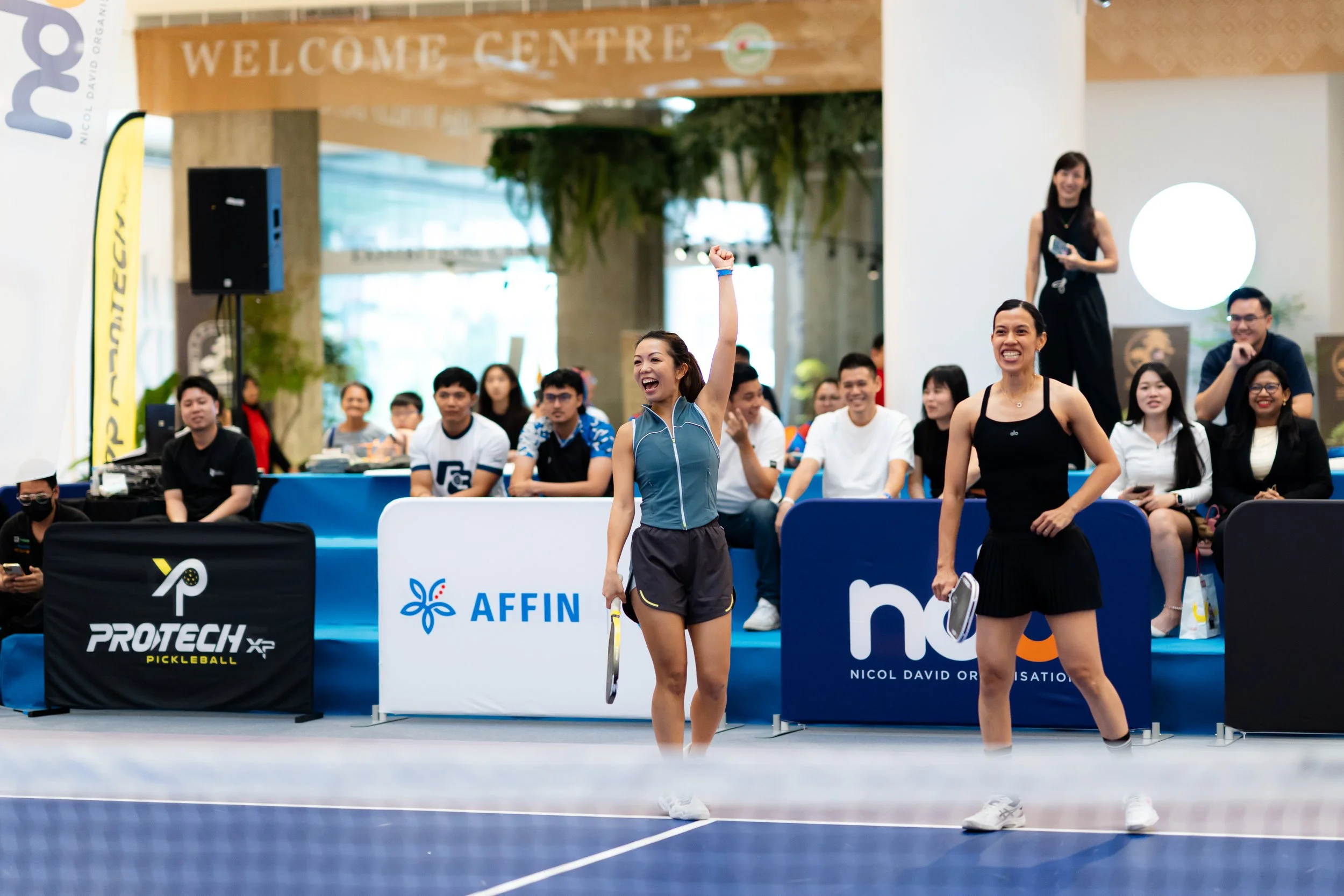 A group of people at a pickleball event with two women in the foreground smiling and holding paddles. The background shows a gathering of seated spectators and banners with logos.