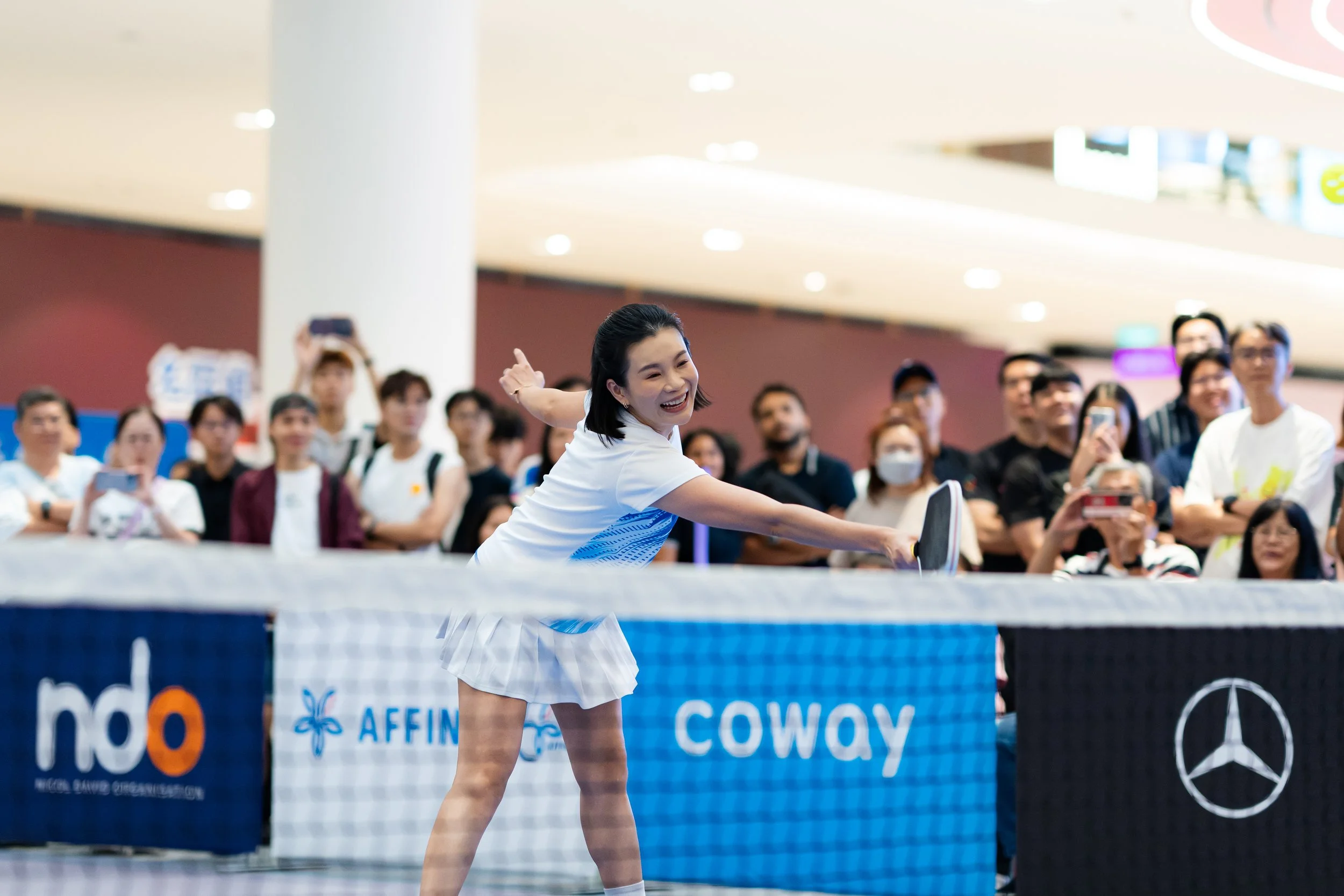 A woman playing table tennis indoors, with a crowd of spectators watching and cheering in the background.