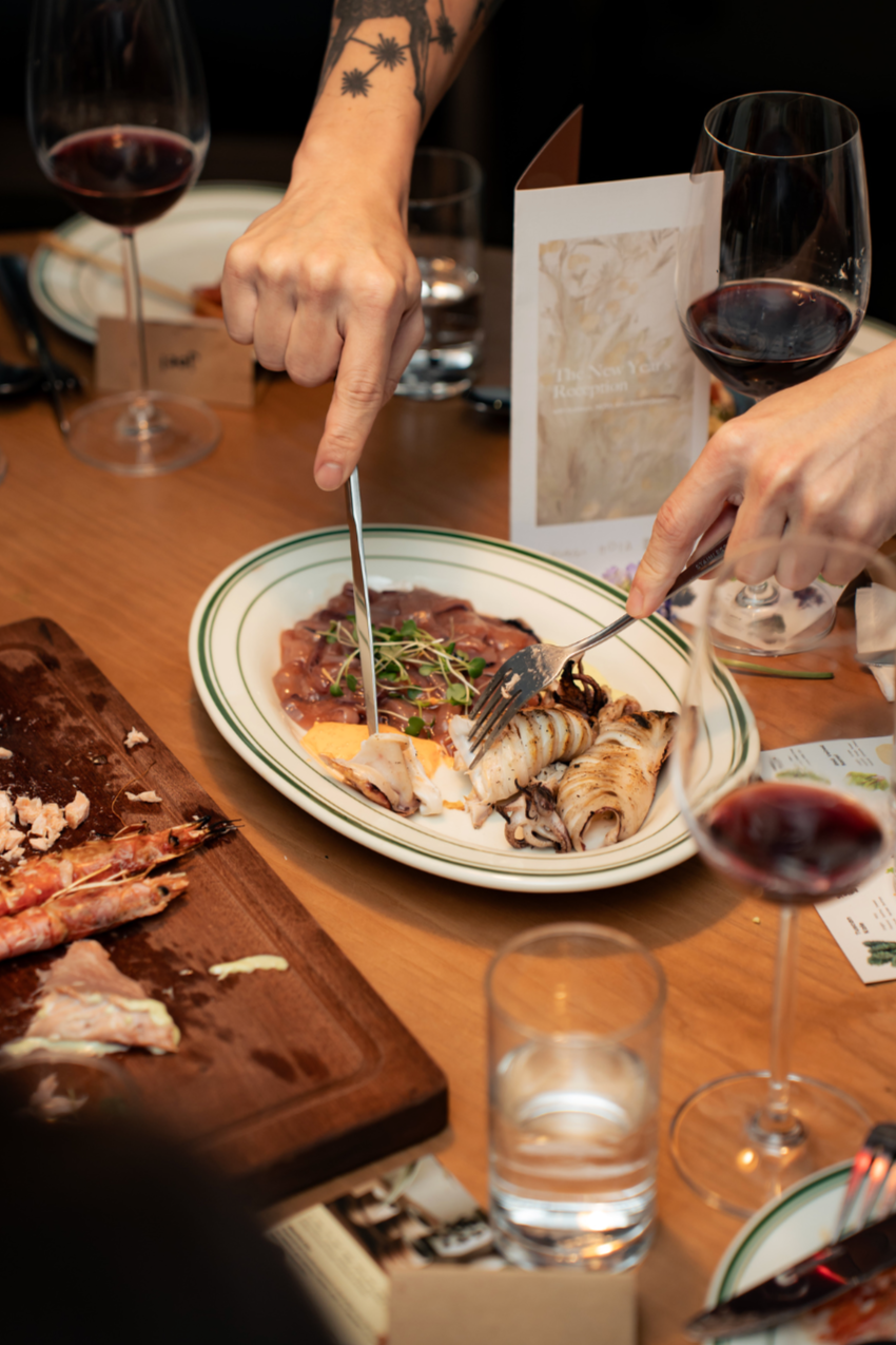 Person using fork and knife to cut into a plate of assorted seafood including squid and fish, with glasses of red wine on a wooden table.