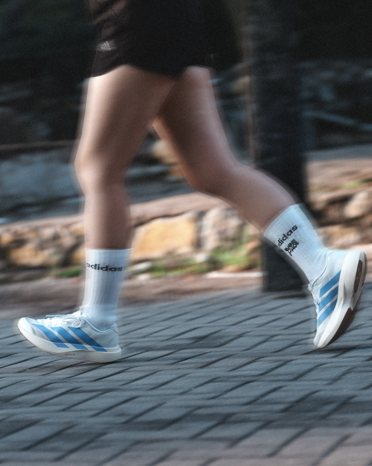 Close-up of a person running on a paved path, wearing white Adidas socks with black text, white and blue athletic shoes, and black shorts, with motion blur indicating movement.