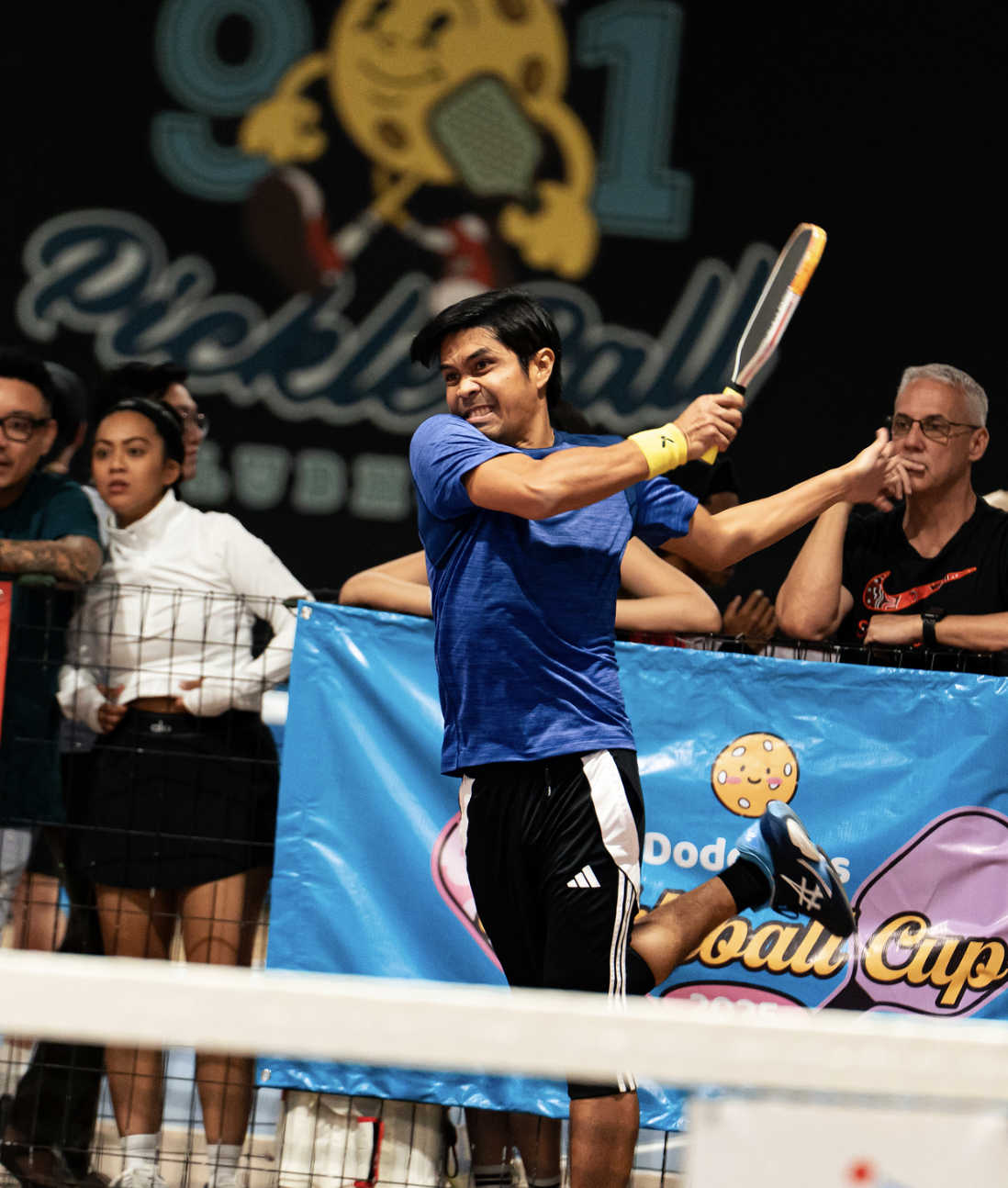 A young man in a blue shirt and black shorts playing pickleball, preparing to hit the ball with a paddle, surrounded by spectators behind a blue barrier at a pickleball tournament.