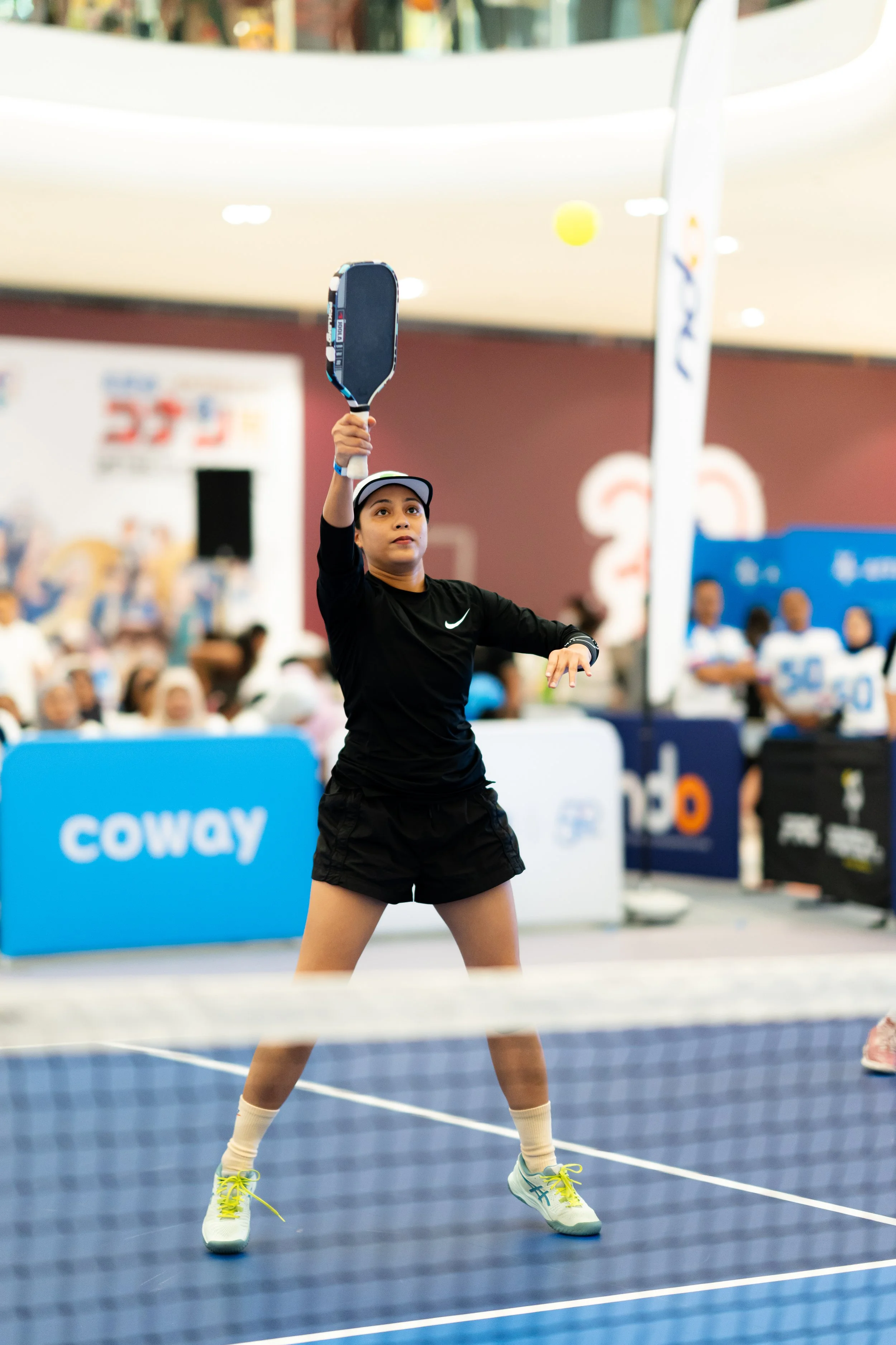A young female athlete in black sportswear playing tennis indoors, preparing to serve with a tennis racket raised in her right hand, on a blue tennis court, with a blurred crowd and signage in the background.