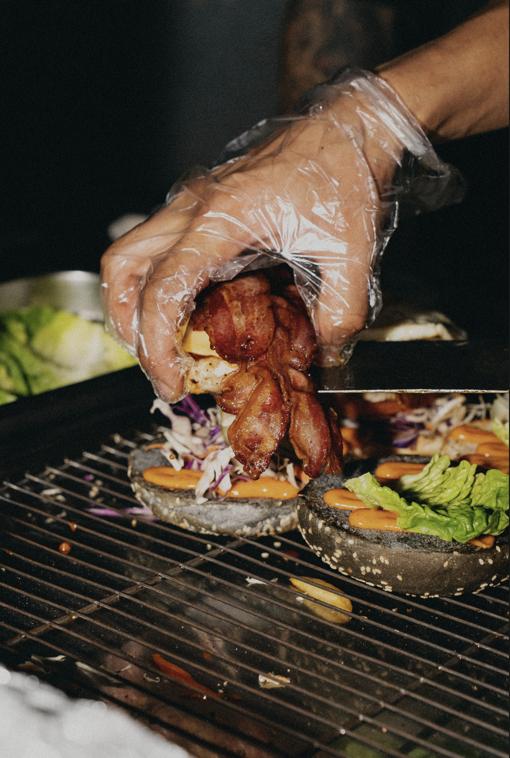 A person wearing a plastic glove is placing crispy bacon on a black sesame seed bun with vegetables for a sandwich, on a grill or cooking surface.