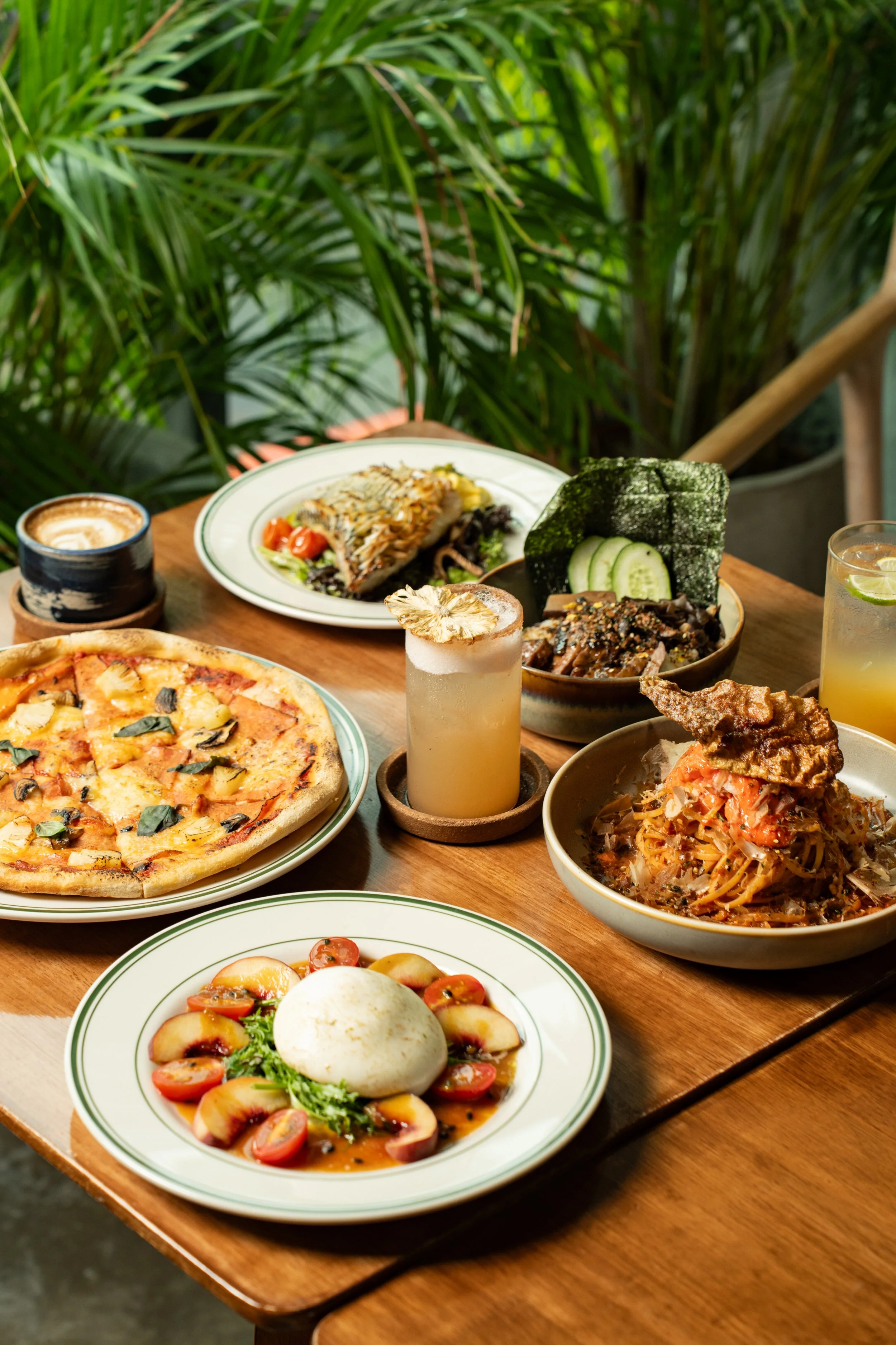 Various plates of food on a wooden table, including pizza, pasta, salad, and drinks, with a background of green plants.