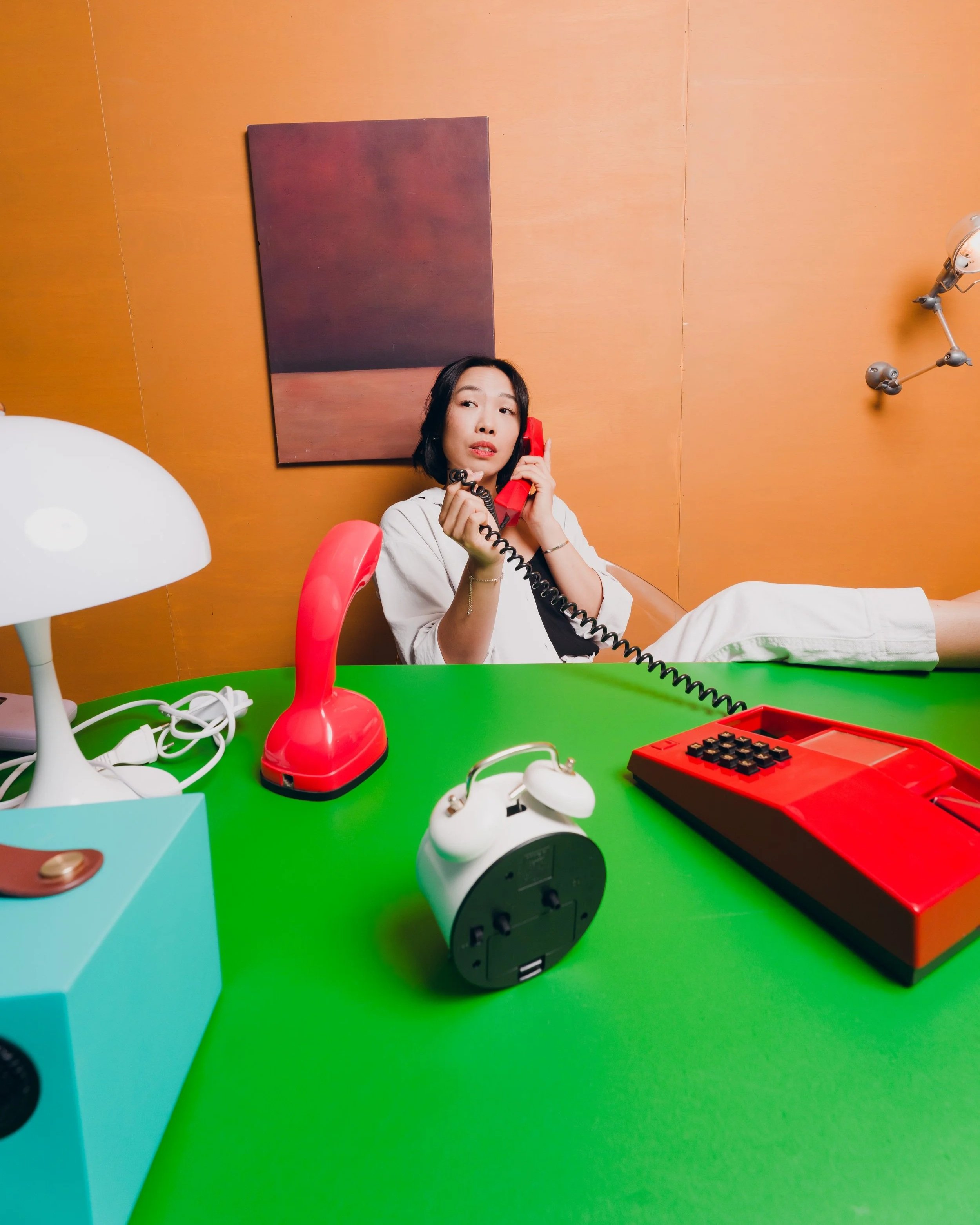 Woman sitting at a brightly colored green table with a red telephone and various vintage electronic devices, speaking on the phone in a room with orange walls and a black rectangular painting.