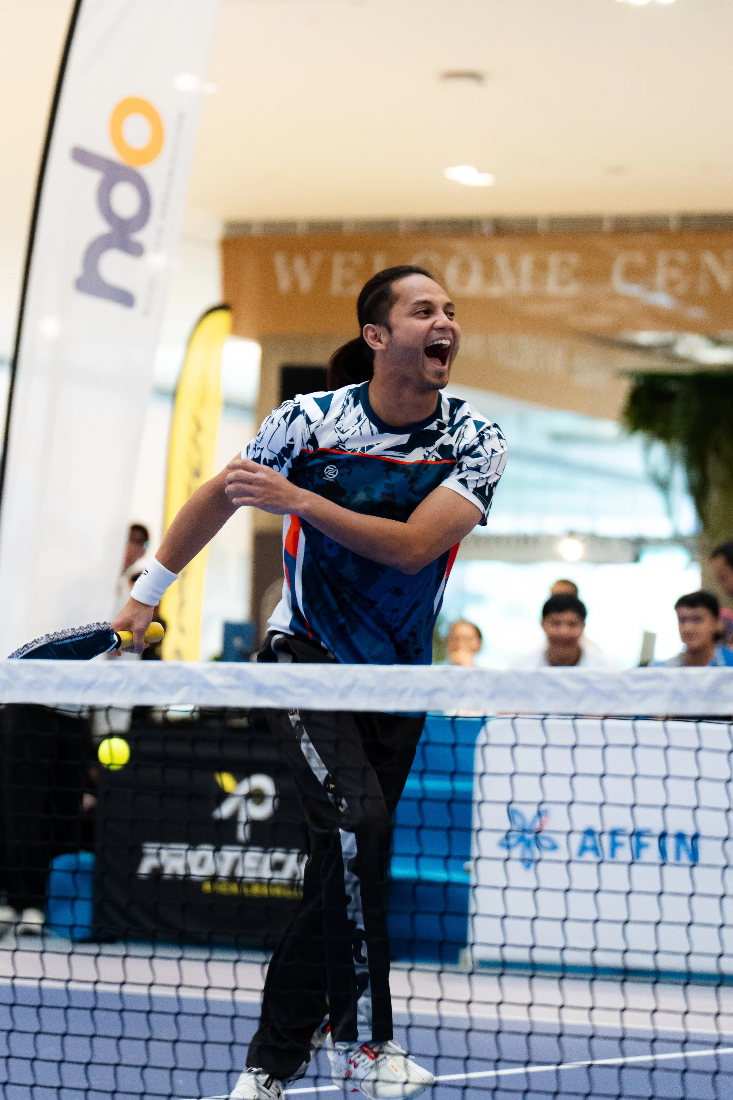 A man in athletic clothing is celebrating by a ping pong table, with his mouth open and arm outstretched, in a public indoor space with spectators and banners.