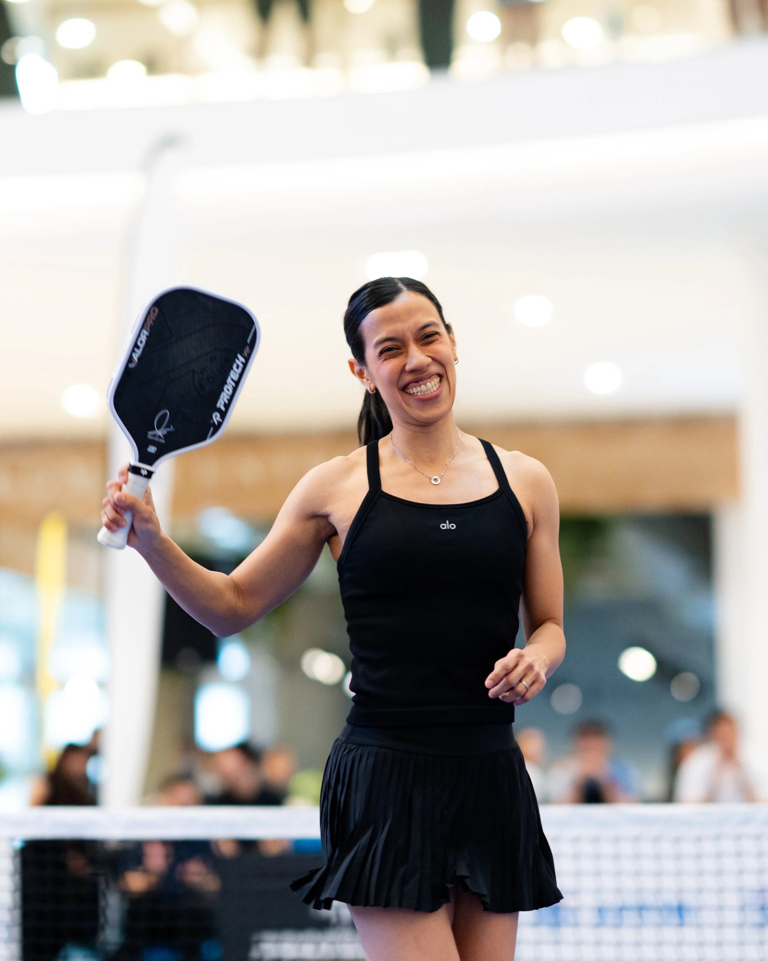 A smiling woman holding a tennis racket, wearing a black athletic top and a pleated black skirt, standing in an indoor sports facility with a blurred background of people and bright lighting.