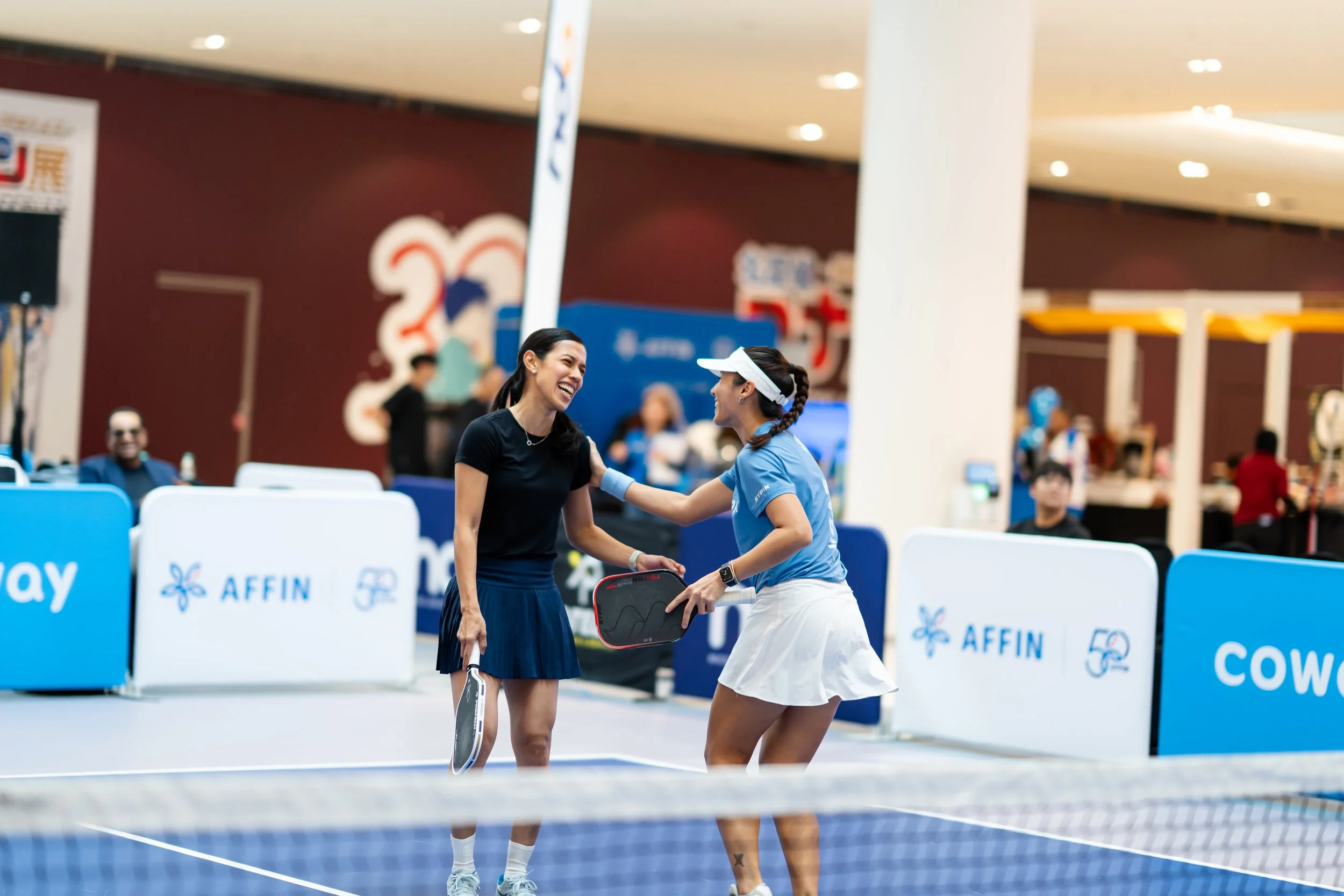 Two female tennis players smiling and shaking hands at a tennis court after a match, with a referee and other spectators in the background.