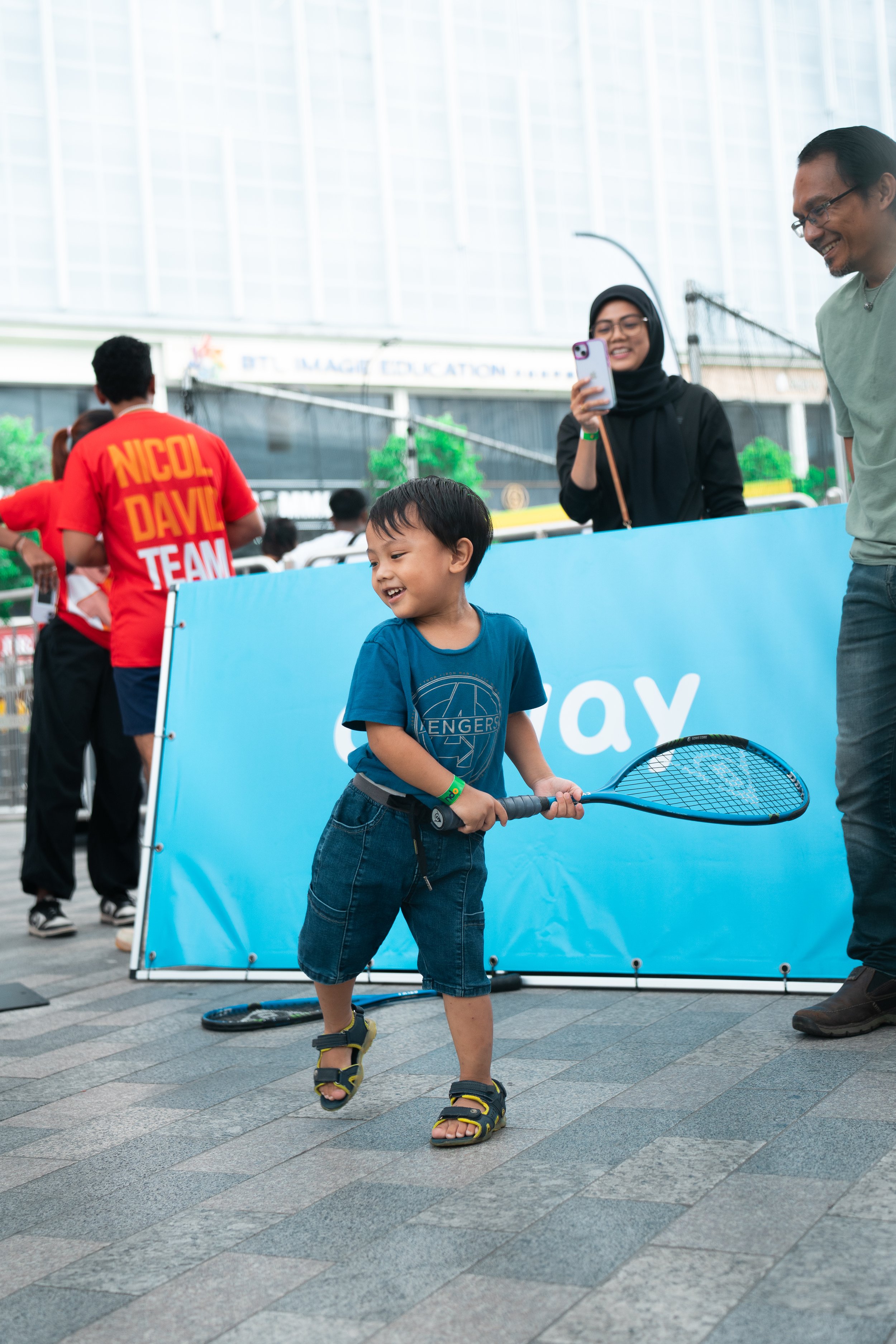 Young boy in sandals and shorts holding a tennis racket, smiling and playing outdoors with a crowd and a blue barrier in the background.