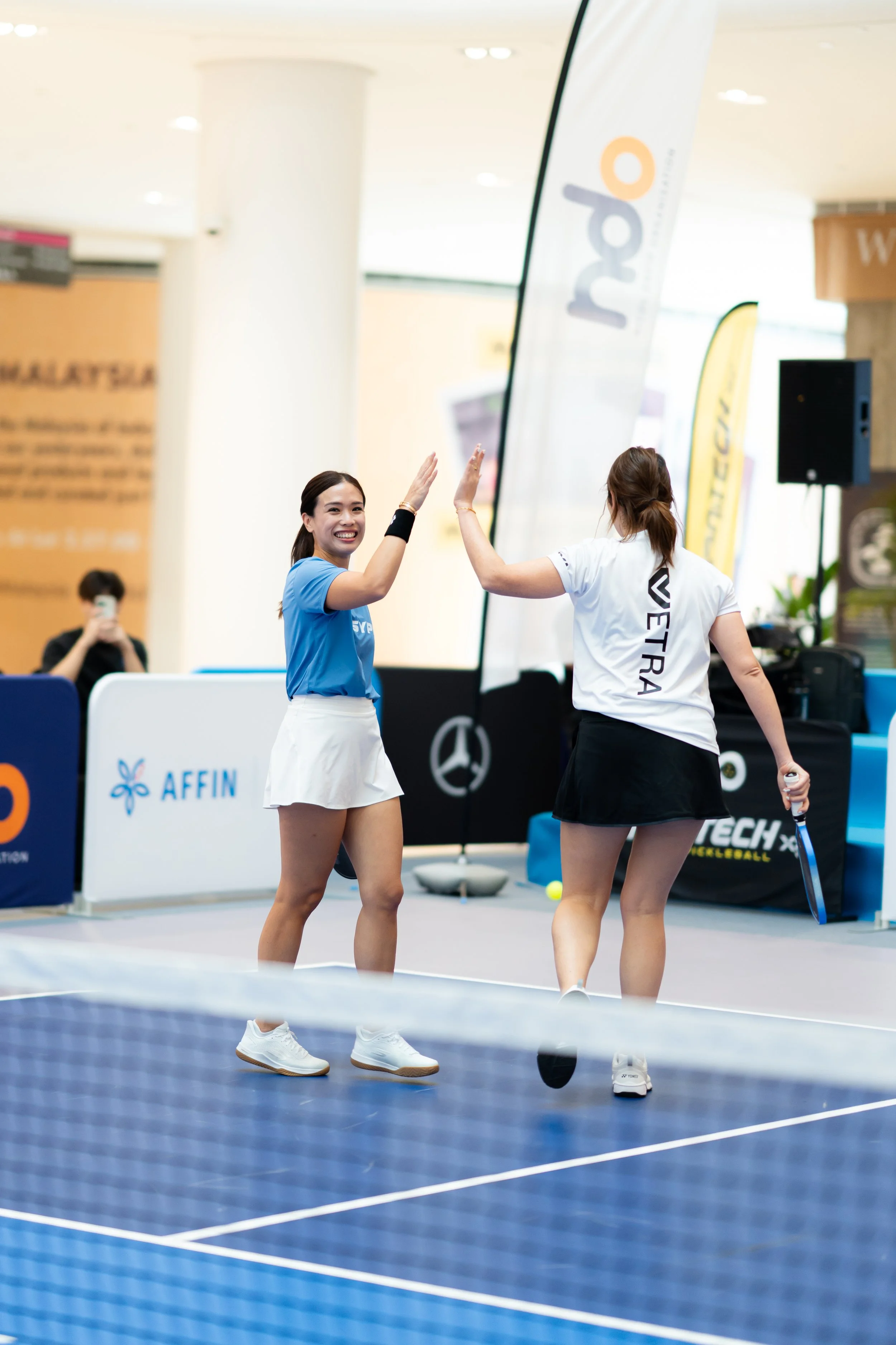 Two women high-fiving on a tennis court with a net in the foreground. One woman is wearing a blue top and white skirt, while the other is wearing a white shirt and black skirt. There are banners and equipment in the background.
