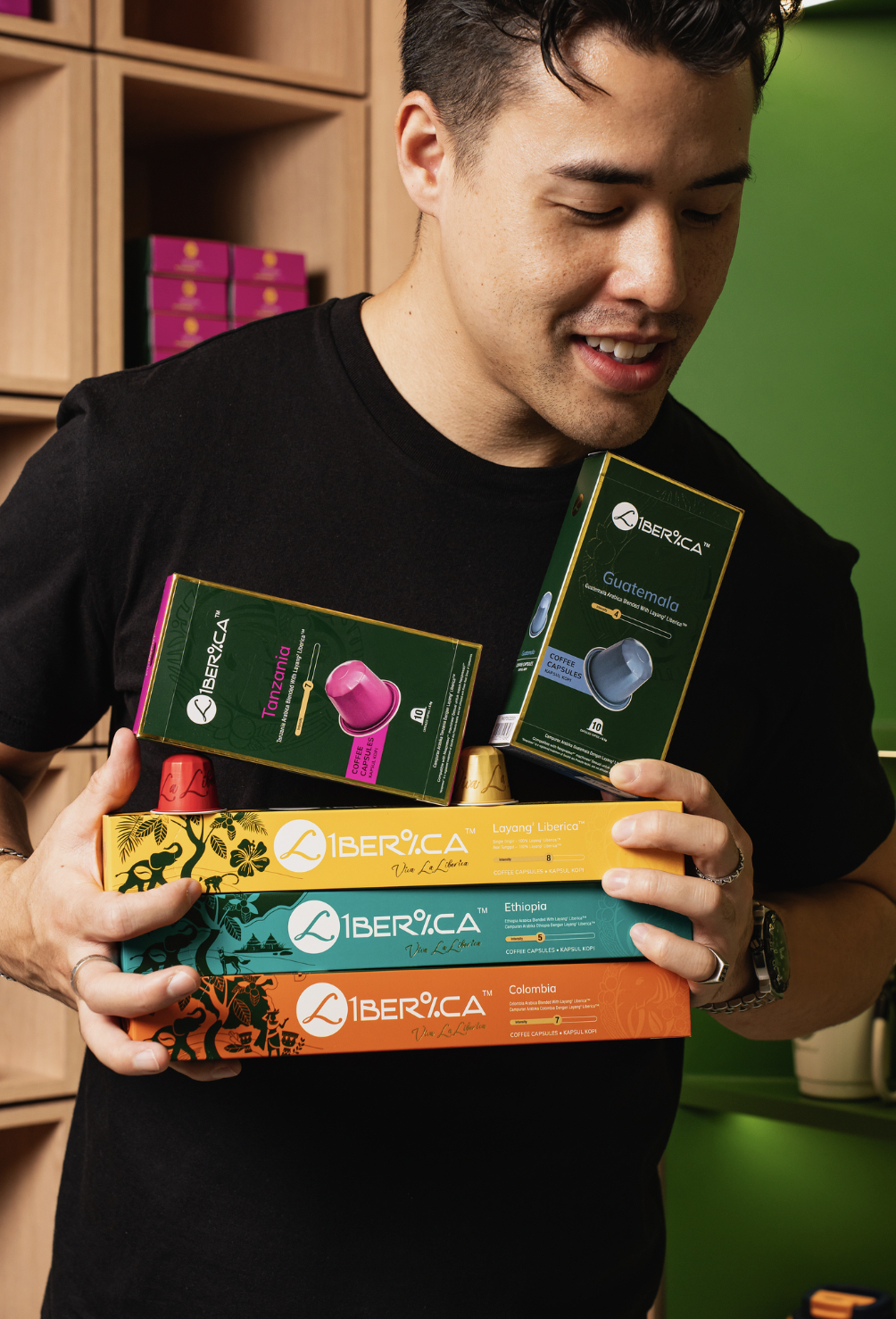 A man holds multiple boxes of Iberica coffee capsules, with some boxes stacked in his arms and others balanced on his shoulder. The boxes are colorful, featuring Guatemala, Ethiopia, Colombia, and Tanzania varieties. He is smiling, wearing a black shirt, and is in a setting with wooden shelves and a green wall in the background.