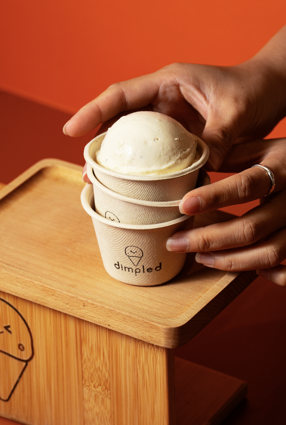 Person holding a cup of vanilla ice cream with a wooden tray and orange background.
