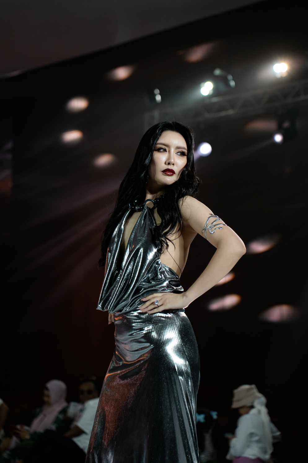 Model with long black hair wearing metallic silver dress, walking on runway with audience in background at fashion show.