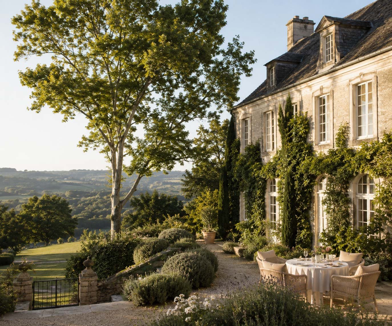 A large stone house covered in greenery with garden dining setup, surrounded by trees and scenic hilly landscape in the background during daytime.