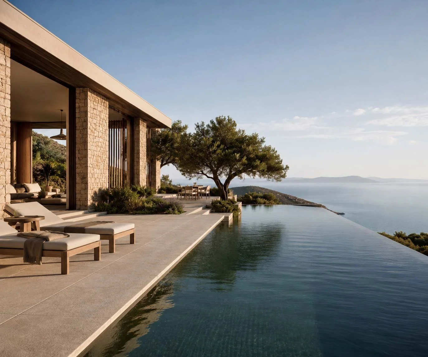 Luxury house with stone walls by an infinity pool overlooking the ocean, with outdoor seating and a tree providing shade.