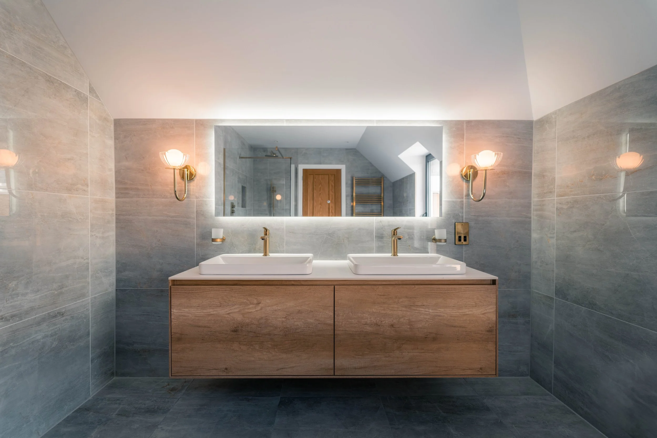 Modern bathroom with dual sinks, large mirror, wall-mounted sconces, wooden cabinetry, and tiled walls and floors.