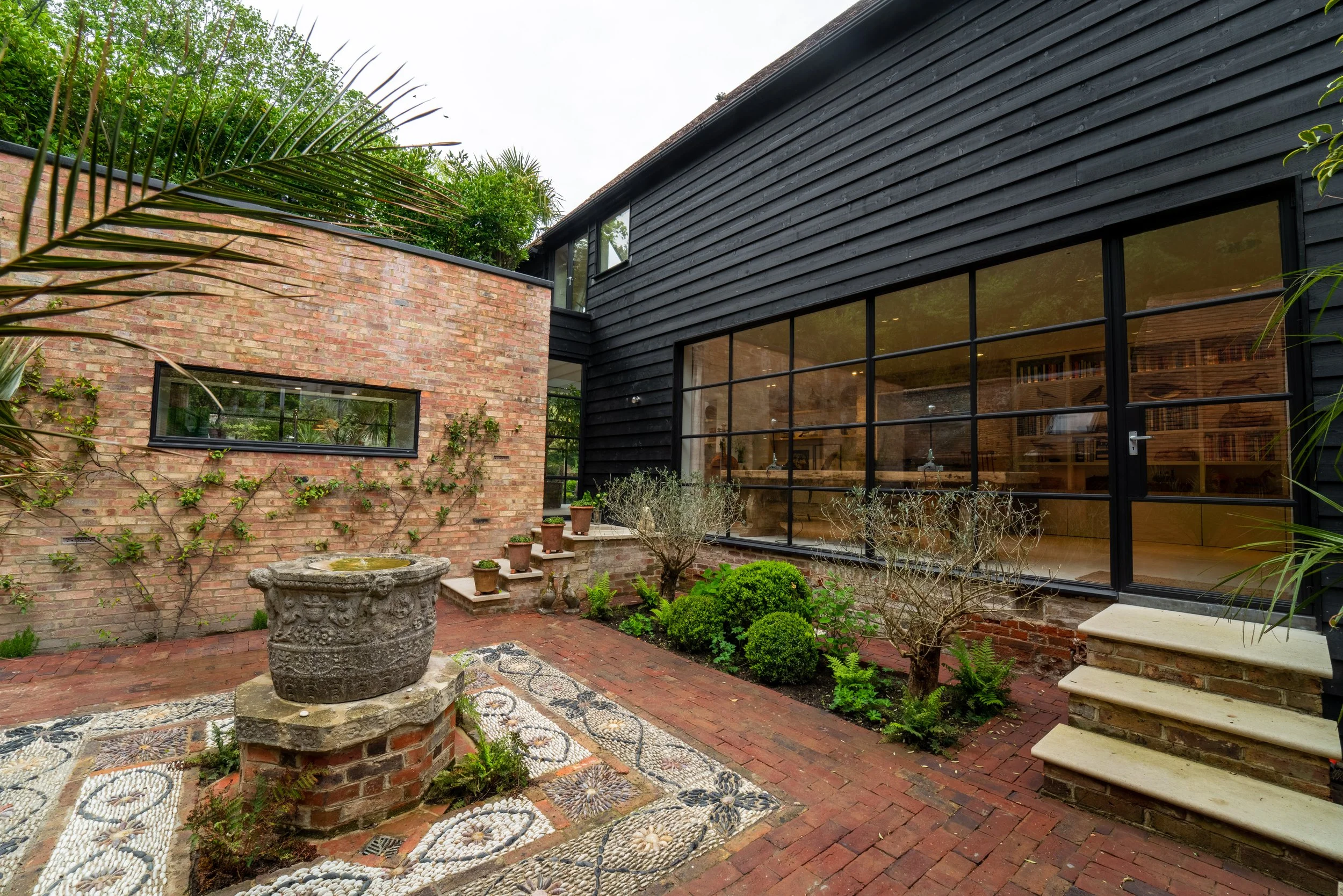 A modern backyard courtyard with brick flooring, a stone planter, potted plants, a black wooden house with large glass doors, greenery, and a staircase.