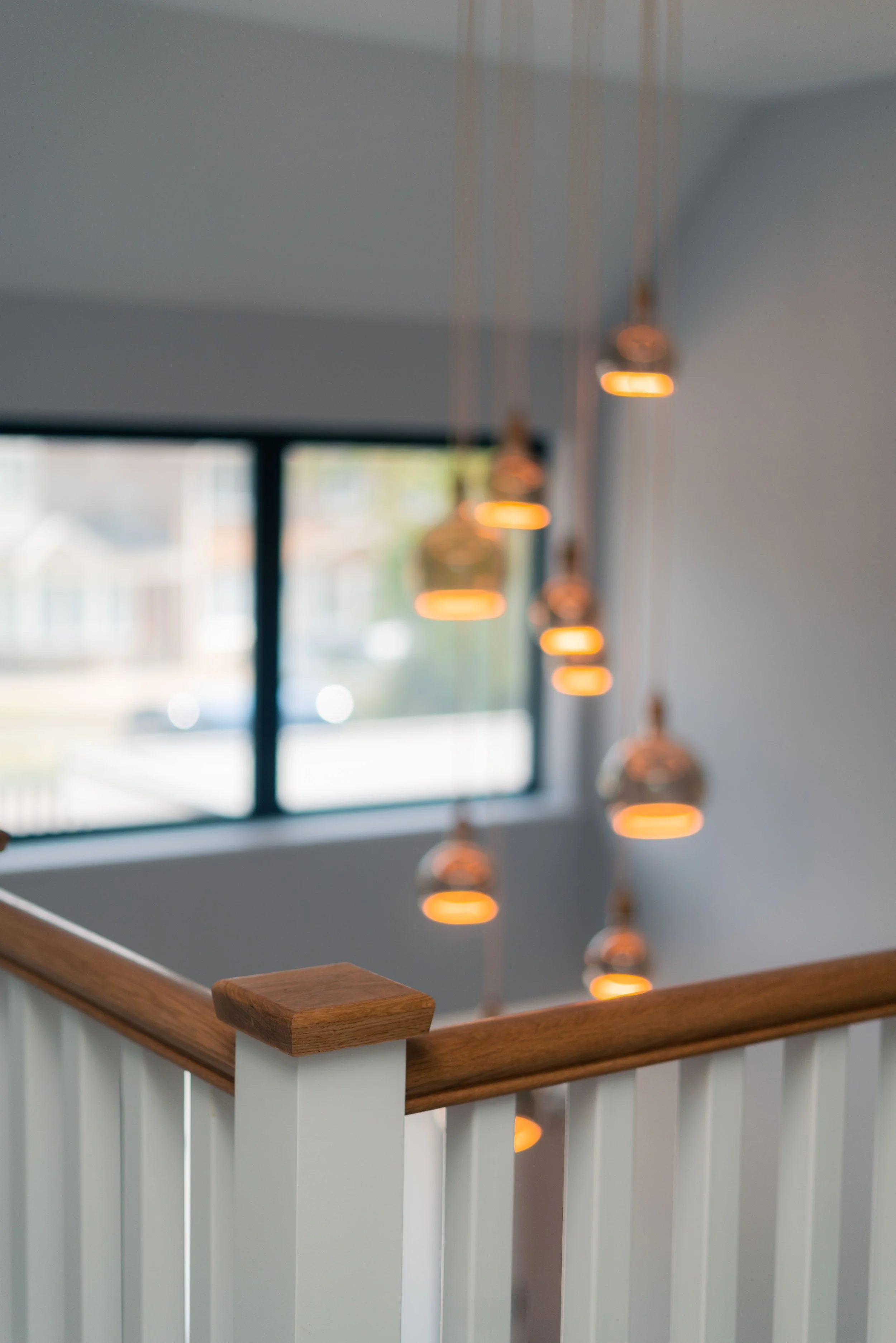 A wooden handrail on a banister with white balusters, overlooking a room with a window and hanging pendant lights.