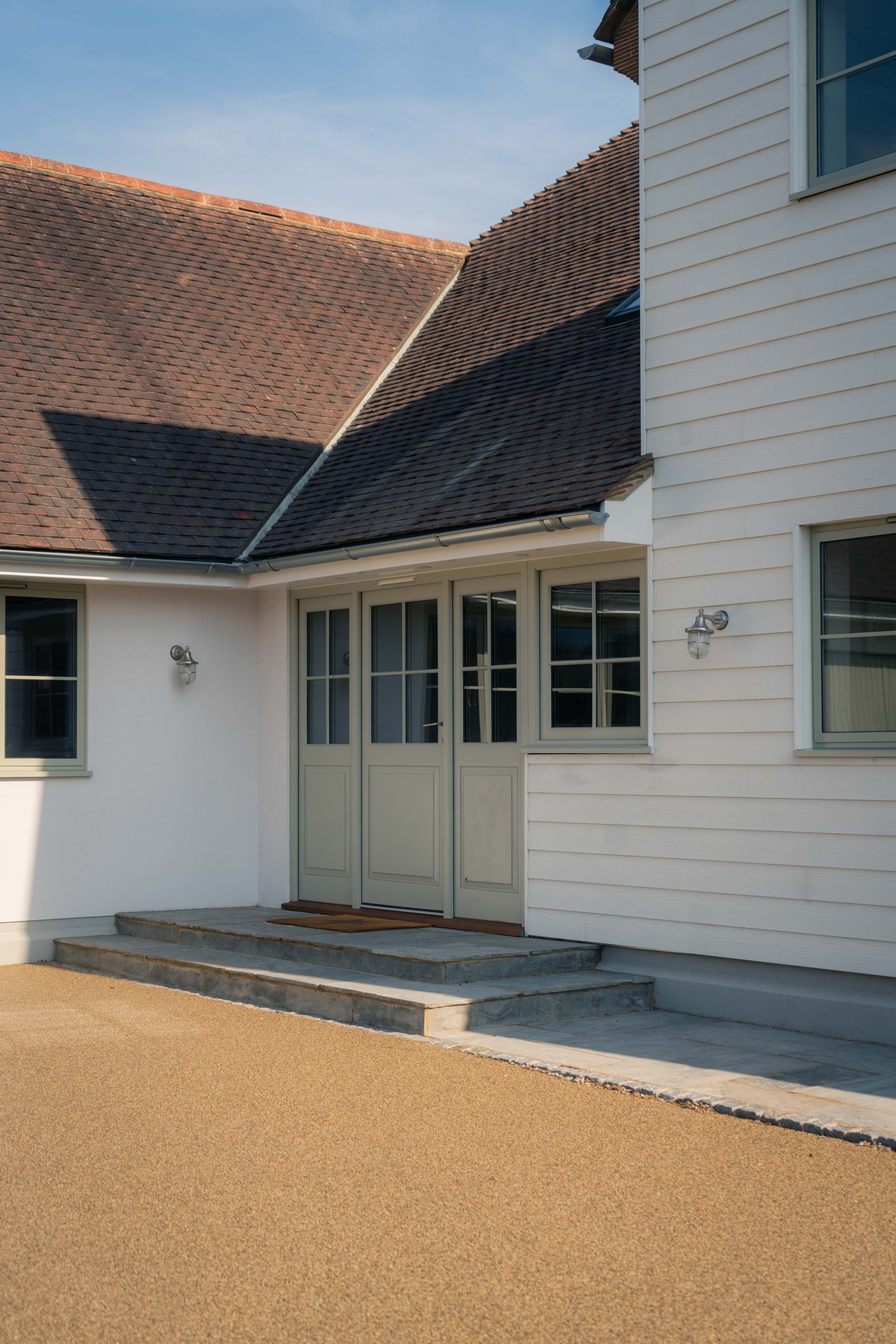 The exterior of a house with a red tile roof, white siding, and multiple windows. The house has a small front porch with steps leading to the entrance, and there are two outdoor wall lights. The sky is clear and blue.