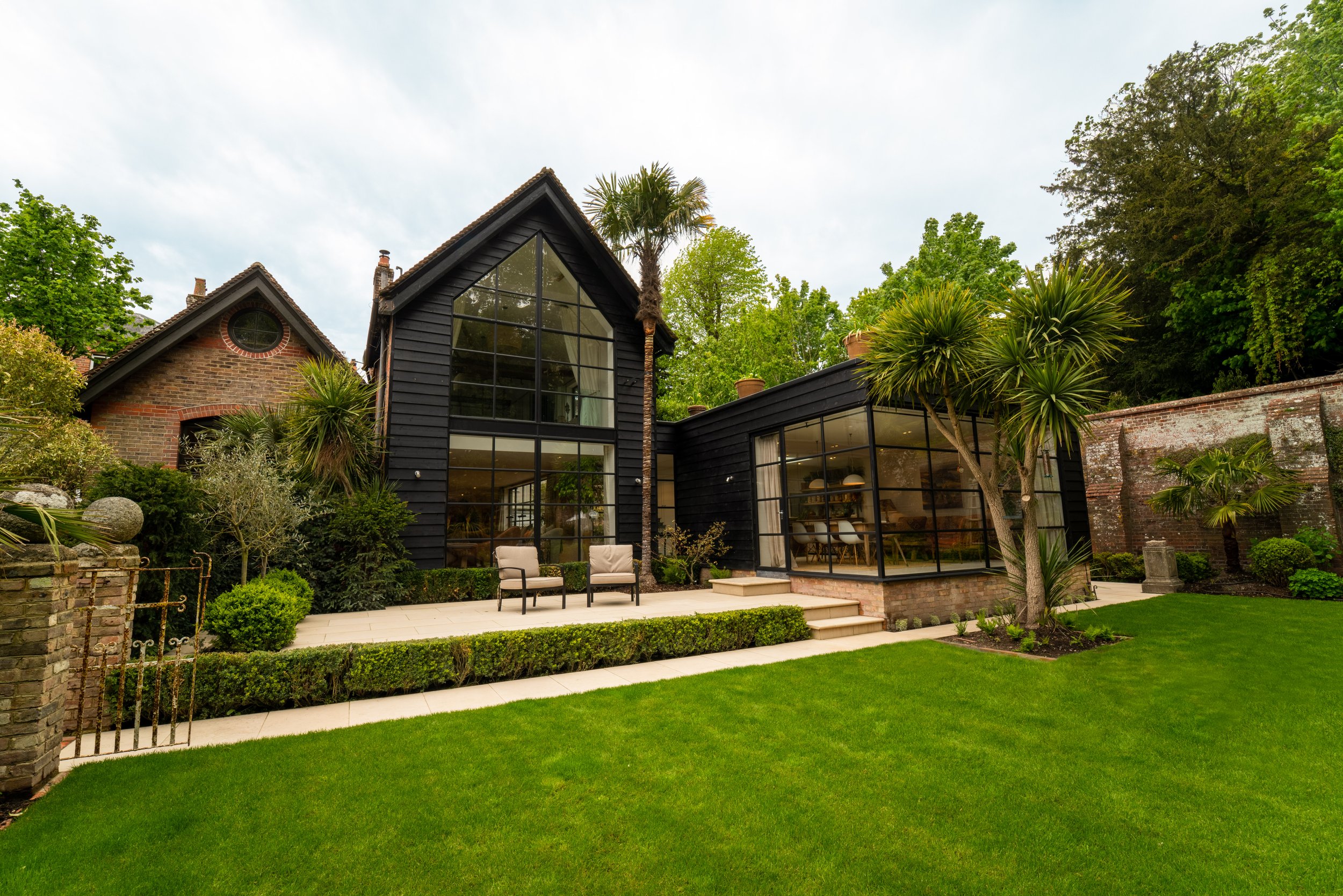 Modern house with large glass window, outdoor seating area, lawn, trees, and brick wall.